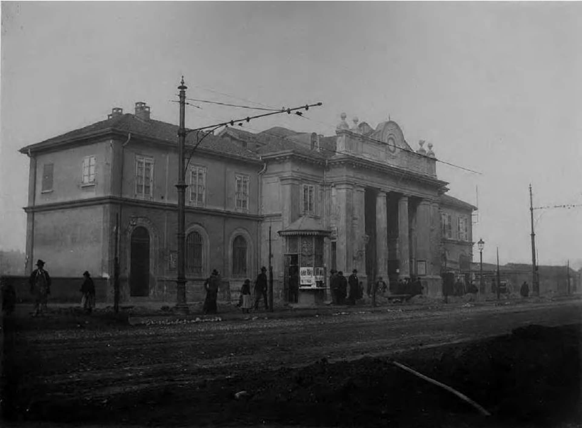 Fotografia storica in bianco e nero dell’edificio della stazione della ferrovia Torino–Ciriè–Lanzo, ripresa da posizione frontale lungo il fronte stradale, che documenta una fase successiva rispetto all’immagine precedente e mostra l’architettura con maggiore definizione e dettaglio. Il fabbricato presenta una composizione simmetrica con corpo centrale monumentale affiancato da due ali laterali a due piani, coperte da tetto a falde e caratterizzate da aperture regolari con cornici modanate. L’ingresso principale è sottolineato da un avancorpo con portico tetrastilo sorretto da colonne di ordine classico, sormontato da un frontone decorato con elementi plastici e timpano curvilineo, che conferisce all’edificio un carattere rappresentativo tipico delle architetture ferroviarie di fine Ottocento. Il piano terra è scandito da aperture ad arco, mentre il piano superiore presenta finestre rettangolari allineate secondo uno schema rigoroso e simmetrico. In primo piano è visibile la strada antistante la stazione, con pavimentazione irregolare, pali della linea tranviaria e alcune persone in attesa o in movimento, elementi che testimoniano l’intenso rapporto tra l’edificio e lo spazio pubblico urbano. La presenza dei cavi aerei e dei lampioni evidenzia l’inserimento della stazione all’interno del sistema di trasporto cittadino, confermandone il ruolo di nodo infrastrutturale e punto di accesso alla città. L’immagine assume valore documentario per la ricostruzione storica del complesso, poiché consente di riconoscere con precisione i caratteri originari del prospetto principale, la configurazione dello spazio antistante e l’aspetto dell’edificio prima delle trasformazioni avvenute nel corso del Novecento.