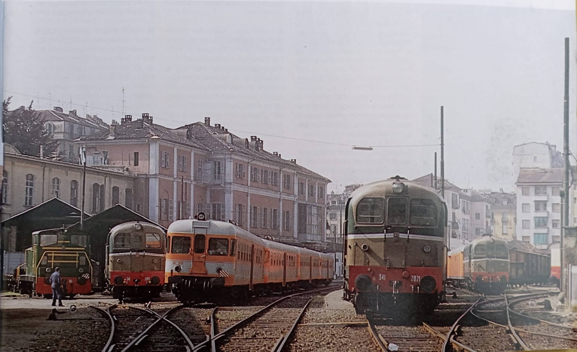 Fotografia storica a colori dell’area ferroviaria adiacente alla stazione Torino–Ciriè–Lanzo, che documenta una fase di utilizzo del complesso come nodo operativo e deposito rotabili. L’immagine è ripresa dall’interno del piazzale ferroviario e mostra diversi convogli fermi sui binari di manovra, evidenziando la configurazione dell’infrastruttura e il rapporto diretto tra gli edifici della stazione e il sistema dei tracciati. In primo piano sono visibili più locomotive e automotrici disposte su binari divergenti, che convergono verso il fascio centrale, definendo una geometria complessa di scambi e raccordi tipica delle aree di deposito e manutenzione. I rotabili, appartenenti a diverse epoche e tipologie, testimoniano l’uso prolungato dell’impianto ferroviario e la sua funzione di capolinea e punto di servizio per la linea. Sullo sfondo si riconosce il fronte edilizio storico della stazione, con fabbricati a più piani caratterizzati da facciate intonacate, finestre regolari e coperture a falde, affiancati da strutture più basse destinate a funzioni tecniche e magazzini. La vicinanza tra edifici urbani e area ferroviaria evidenzia l’integrazione originaria tra infrastruttura e tessuto cittadino, tipica delle stazioni di fine Ottocento e primo Novecento. L’immagine ha valore documentario per la ricostruzione storica del sito, poiché mostra l’assetto del piazzale ferroviario prima delle trasformazioni successive e permette di comprendere l’estensione delle aree di servizio, la disposizione dei binari e il ruolo del complesso come infrastruttura ferroviaria attiva all’interno della città.