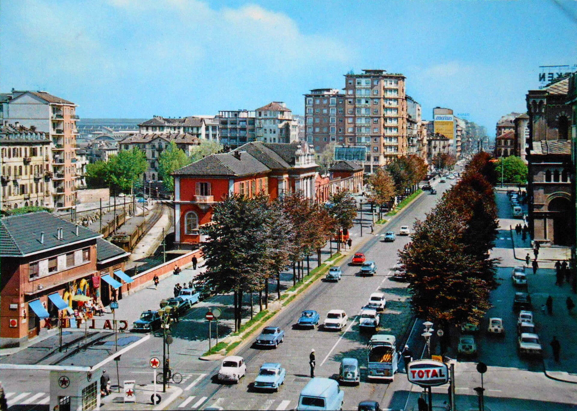 Fotografia storica a colori dell’area urbana di Corso Giulio Cesare a Torino, con vista prospettica verso il complesso della stazione della linea Torino–Ciriè–Lanzo, ripresa da posizione sopraelevata lungo l’asse stradale principale. L’immagine documenta la configurazione del quartiere in una fase di pieno utilizzo dell’infrastruttura ferroviaria, evidenziando il rapporto diretto tra la stazione, il tessuto residenziale circostante e la viabilità urbana. Al centro della composizione è riconoscibile l’edificio storico della stazione, con facciate intonacate di colore rosso e corpo centrale monumentale con frontone decorato, affacciato sul viale alberato. Accanto al fabbricato principale sono visibili i binari e le pensiline della linea ferroviaria, che scorrono parallelamente alla strada, confermando la presenza dell’impianto come infrastruttura integrata nel sistema urbano. Il corso è caratterizzato da una sezione ampia con doppia carreggiata, spartitraffico alberato e traffico veicolare intenso, elementi che testimoniano la trasformazione del quartiere in asse urbano di scorrimento. Ai lati della strada si sviluppano edifici residenziali multipiano realizzati nel secondo dopoguerra, che definiscono un fronte edilizio continuo e più alto rispetto al fabbricato storico della stazione, evidenziando il progressivo processo di densificazione della zona. In primo piano sono presenti attività commerciali, distributore di carburante, parcheggi e attraversamenti pedonali, elementi che contribuiscono a descrivere il contesto urbano degli anni del boom economico e il ruolo del complesso ferroviario come elemento ancora attivo ma già inserito in un sistema cittadino in forte trasformazione. L’immagine ha valore di documento storico-urbanistico, poiché consente di comprendere l’evoluzione dell’intorno costruito, il rapporto tra infrastruttura ferroviaria e città e la progressiva modifica del paesaggio urbano attorno all’edificio oggetto di intervento.