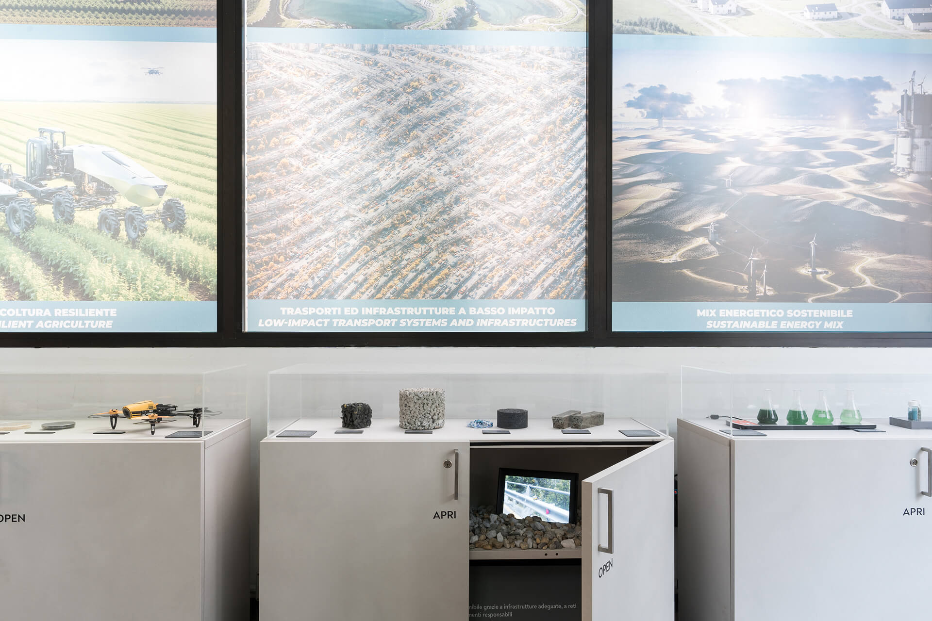 Allestimento espositivo interno costituito da una serie di teche museali con struttura in pannelli bianchi e copertura trasparente, contenenti campioni di materiali, modelli e dispositivi dimostrativi legati ai temi della sostenibilità ambientale e dell’innovazione tecnologica. All’interno delle vetrine sono visibili campioni inerti, materiali compositi, modelli in scala e strumenti scientifici, disposti su supporti espositivi con etichette descrittive. Sullo sfondo, grandi pannelli grafici retroilluminati illustrano scenari legati alla transizione ecologica, tra cui agricoltura resiliente, sistemi di trasporto a basso impatto e mix energetico sostenibile, con immagini di infrastrutture, paesaggi produttivi e impianti per energie rinnovabili. Una delle teche presenta uno sportello apribile con indicazione bilingue “APRI / OPEN”, che consente l’interazione diretta con il contenuto espositivo, evidenziando il carattere didattico e divulgativo dell’allestimento. L’immagine documenta uno spazio museale o centro di ricerca allestito con finalità informative, in cui elementi espositivi, grafica e dispositivi interattivi sono integrati in un sistema unitario di comunicazione scientifica.