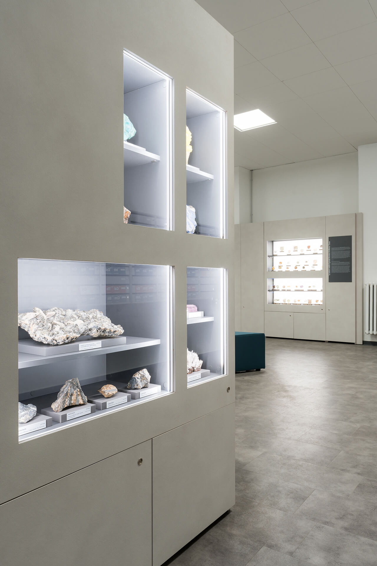 Vista interna di ambiente museale con pareti attrezzate modulari destinate all’esposizione di campioni mineralogici, realizzate mediante pannellature continue di colore chiaro nelle quali sono ricavate nicchie illuminate incassate. Le teche, di diverse dimensioni e disposte su più livelli, contengono rocce e minerali classificati, collocati su supporti individuali con etichette identificative e organizzati secondo criteri scientifici. L’illuminazione integrata perimetrale garantisce una luce diffusa e uniforme all’interno dei vani, evidenziando le caratteristiche morfologiche e cromatiche dei campioni. In primo piano è visibile un modulo espositivo angolare che definisce la scansione dello spazio e introduce la sequenza delle pareti attrezzate, mentre sullo sfondo si intravede un ulteriore sistema di teche illuminate inserito lungo la parete opposta. Il soffitto è costituito da controsoffitto modulare con corpi illuminanti incassati, mentre la pavimentazione continua in materiale resiliente di tonalità neutra contribuisce alla percezione di ordine e continuità dell’ambiente. L’immagine documenta un allestimento museale permanente progettato per la divulgazione scientifica, caratterizzato da un sistema espositivo modulare integrato che unisce arredi, illuminazione e apparati informativi in un unico dispositivo coerente, pensato per garantire leggibilità, conservazione dei reperti e facilità di fruizione da parte dei visitatori.