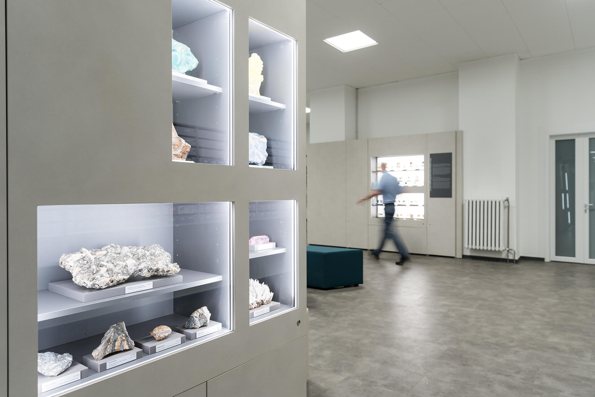Vista interna di ambiente museale con pareti attrezzate modulari destinate all’esposizione di campioni mineralogici, realizzate mediante pannellature continue nelle quali sono ricavate nicchie illuminate incassate. In primo piano è visibile un modulo espositivo con teche di diverse dimensioni contenenti rocce e minerali classificati, disposti su supporti individuali con etichette identificative e organizzati secondo criteri scientifici. L’illuminazione integrata perimetrale delle vetrine garantisce una luce uniforme e controllata, idonea alla valorizzazione delle caratteristiche materiche e cromatiche dei campioni. Lo spazio si apre verso un ambiente più ampio, pavimentato con superficie continua in materiale resiliente di tonalità neutra e coperto da controsoffitto modulare con corpi illuminanti incassati. Sullo sfondo è visibile un ulteriore sistema espositivo illuminato, affiancato da pannelli informativi e da elementi di arredo per la sosta dei visitatori. La presenza di una figura in movimento evidenzia la scala dell’ambiente e la modalità di fruizione del percorso museale. L’immagine documenta un allestimento museale permanente progettato per la divulgazione scientifica, caratterizzato da un sistema espositivo modulare integrato che unisce arredi, illuminazione e apparati informativi in un dispositivo unitario, pensato per garantire leggibilità, conservazione dei reperti e continuità percettiva dello spazio espositivo.
