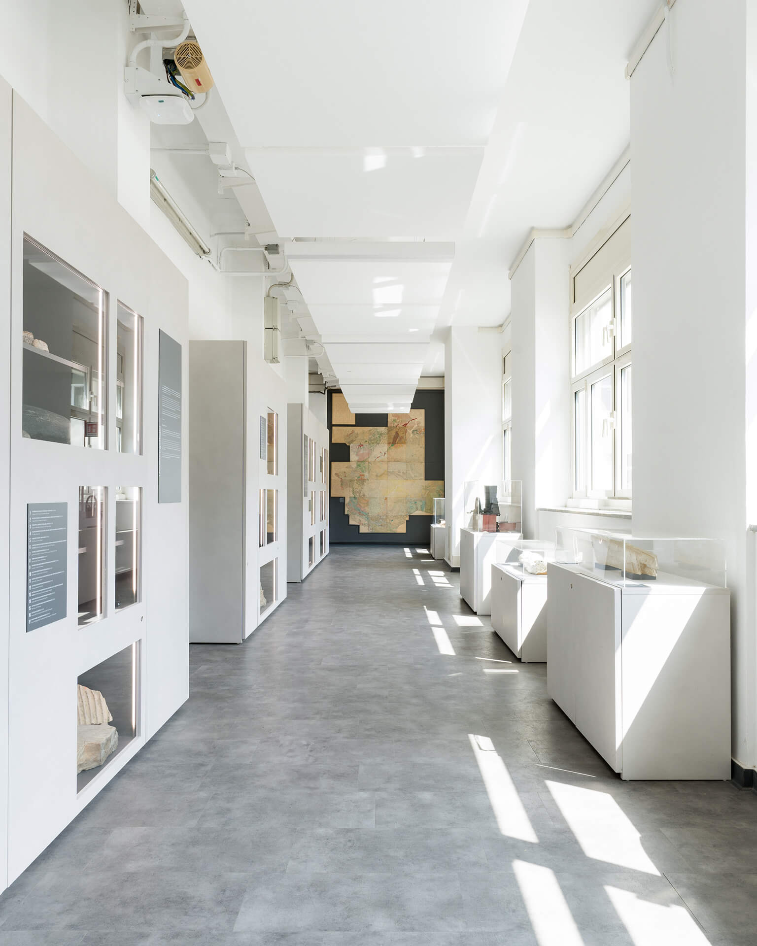 Vista prospettica di corridoio espositivo all’interno di spazio museale, caratterizzato da un percorso longitudinale definito da pareti attrezzate modulari con teche illuminate incassate sul lato sinistro e da una sequenza di basamenti espositivi con vetrine sul lato destro, collocati in corrispondenza delle aperture finestrate. Le nicchie integrate nelle pannellature contengono campioni litologici e reperti scientifici accompagnati da pannelli informativi verticali, inseriti in un sistema grafico coordinato destinato alla divulgazione delle scienze della Terra. L’ambiente è illuminato sia da luce naturale proveniente dalle finestre laterali sia da illuminazione artificiale integrata nel controsoffitto, costituito da elementi ribassati lineari che nascondono impianti e canalizzazioni tecniche, lasciate parzialmente a vista nella fascia superiore. La pavimentazione continua in materiale resiliente di tonalità grigia contribuisce alla lettura uniforme dello spazio e alla continuità del percorso museale. Sul fondo del corridoio è visibile un grande apparato grafico cartografico montato a parete, che funge da elemento conclusivo del percorso espositivo e da supporto didattico. L’immagine documenta un allestimento museale permanente progettato come sistema integrato di arredi, illuminazione e apparati informativi, finalizzato a garantire chiarezza espositiva, conservazione dei reperti e fluidità della fruizione da parte dei visitatori.