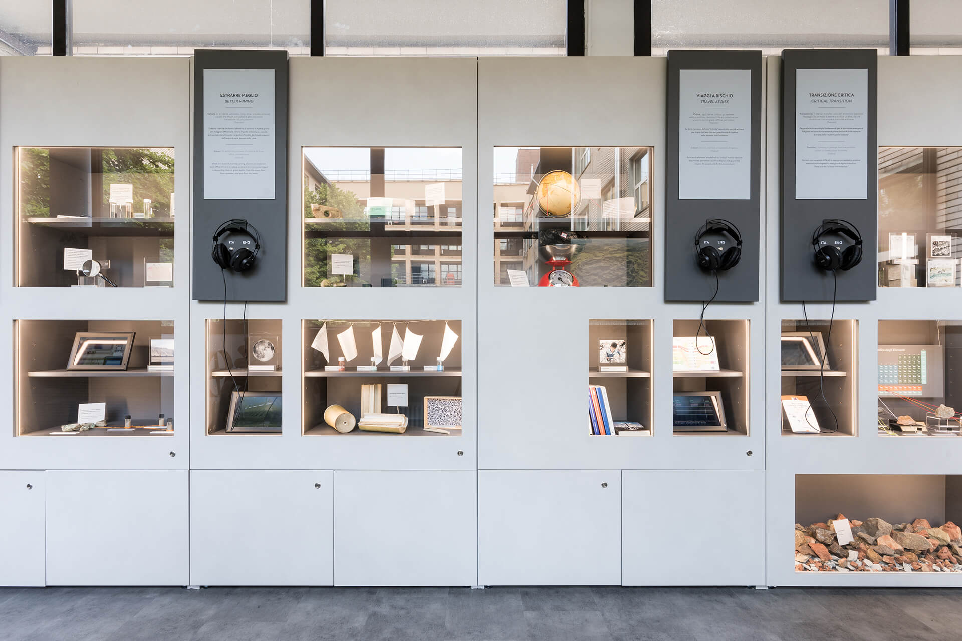 Sistema espositivo modulare costituito da parete attrezzata con pannellature continue in finitura chiara, integrate con nicchie illuminate e vetrine incassate destinate all’esposizione di oggetti, campioni e materiali documentali. All’interno dei vani sono collocati modelli, strumenti scientifici, campioni litici, dispositivi tecnologici e supporti multimediali, organizzati secondo un percorso tematico a carattere didattico-scientifico. Sulla parte superiore della parete sono installati pannelli informativi verticali con testi bilingue e postazioni audio dotate di cuffie, che consentono la fruizione di contenuti sonori di approfondimento. Le teche retroilluminate e i ripiani espositivi sono integrati nella struttura dell’allestimento, mentre nella fascia inferiore sono presenti ante di servizio per l’ispezione e la manutenzione. L’insieme configura un allestimento museale interattivo, progettato per la divulgazione di contenuti legati a scienza, ambiente e innovazione, in cui elementi grafici, supporti audiovisivi e oggetti esposti sono coordinati in un sistema unitario di comunicazione.