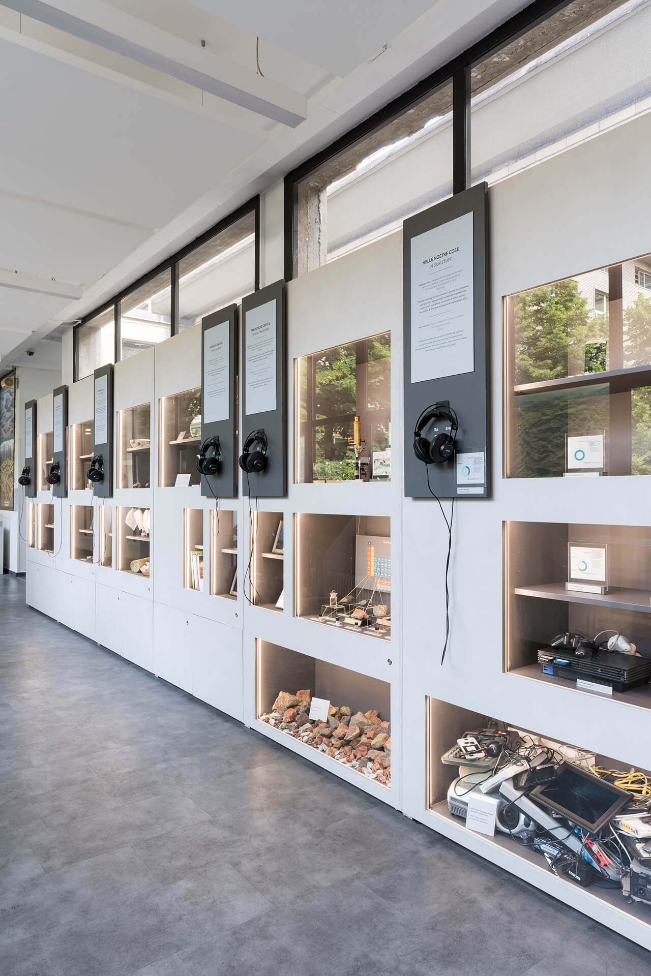 Vista prospettica di parete espositiva all’interno di spazio museale o centro di divulgazione scientifica, costituita da un sistema modulare continuo in pannelli laccati chiari con nicchie illuminate e vetrine incassate destinate alla presentazione di oggetti, campioni e dispositivi tecnologici. Le teche, integrate nella struttura dell’arredo, ospitano materiali litici, strumenti elettronici, modelli, supporti multimediali e documentazione tecnica, organizzati secondo un percorso tematico a carattere didattico. Nella parte superiore sono installati pannelli informativi verticali con testi bilingue e postazioni audio dotate di cuffie per l’ascolto di contenuti di approfondimento, fissate su supporti metallici applicati alla parete. La fascia alta dell’ambiente è caratterizzata da serramenti continui con profili scuri e superfici vetrate opaline che garantiscono illuminazione naturale diffusa, mentre il pavimento in materiale continuo di colore neutro contribuisce alla lettura unitaria dello spazio. L’immagine documenta un allestimento permanente progettato per la comunicazione scientifica e ambientale, in cui elementi grafici, teche interattive e dispositivi audiovisivi sono coordinati in un sistema espositivo integrato.