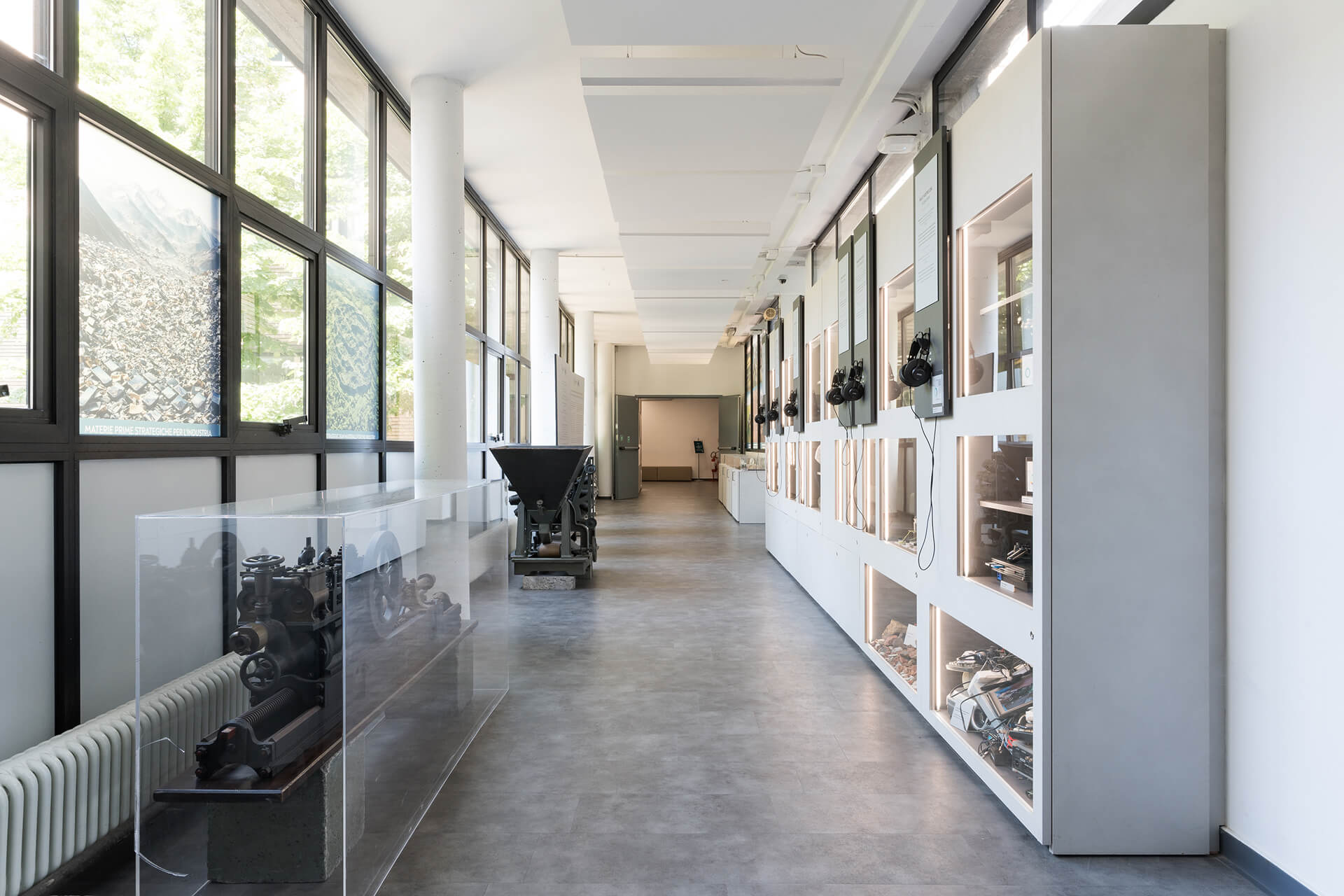 Vista longitudinale di spazio espositivo interno organizzato come corridoio museale, caratterizzato da un sistema continuo di arredi integrati lungo la parete destra, costituiti da moduli espositivi con nicchie illuminate, teche incassate e pannelli informativi verticali dotati di postazioni audio con cuffie per l’ascolto di contenuti multimediali. Le vetrine ospitano campioni, strumenti tecnici, dispositivi elettronici e materiali documentali legati ai temi della ricerca scientifica, dell’industria e dell’innovazione tecnologica, disposti secondo un percorso didattico sequenziale. Sul lato opposto, una serie di grandi serramenti continui con telaio metallico scuro garantisce illuminazione naturale diffusa e mette in relazione lo spazio interno con l’esterno alberato, mentre al centro del percorso sono collocati macchinari storici e apparecchiature industriali protetti da teche trasparenti, utilizzati come elementi espositivi. Il soffitto è configurato con controsoffitti lineari ribassati che integrano l’illuminazione e gli impianti, mentre la pavimentazione continua in materiale resiliente di colore neutro contribuisce alla lettura unitaria dello spazio. L’immagine documenta un allestimento museale permanente, progettato per la divulgazione scientifica e tecnologica, in cui arredi, grafica, dispositivi interattivi e reperti sono coordinati in un sistema espositivo integrato.