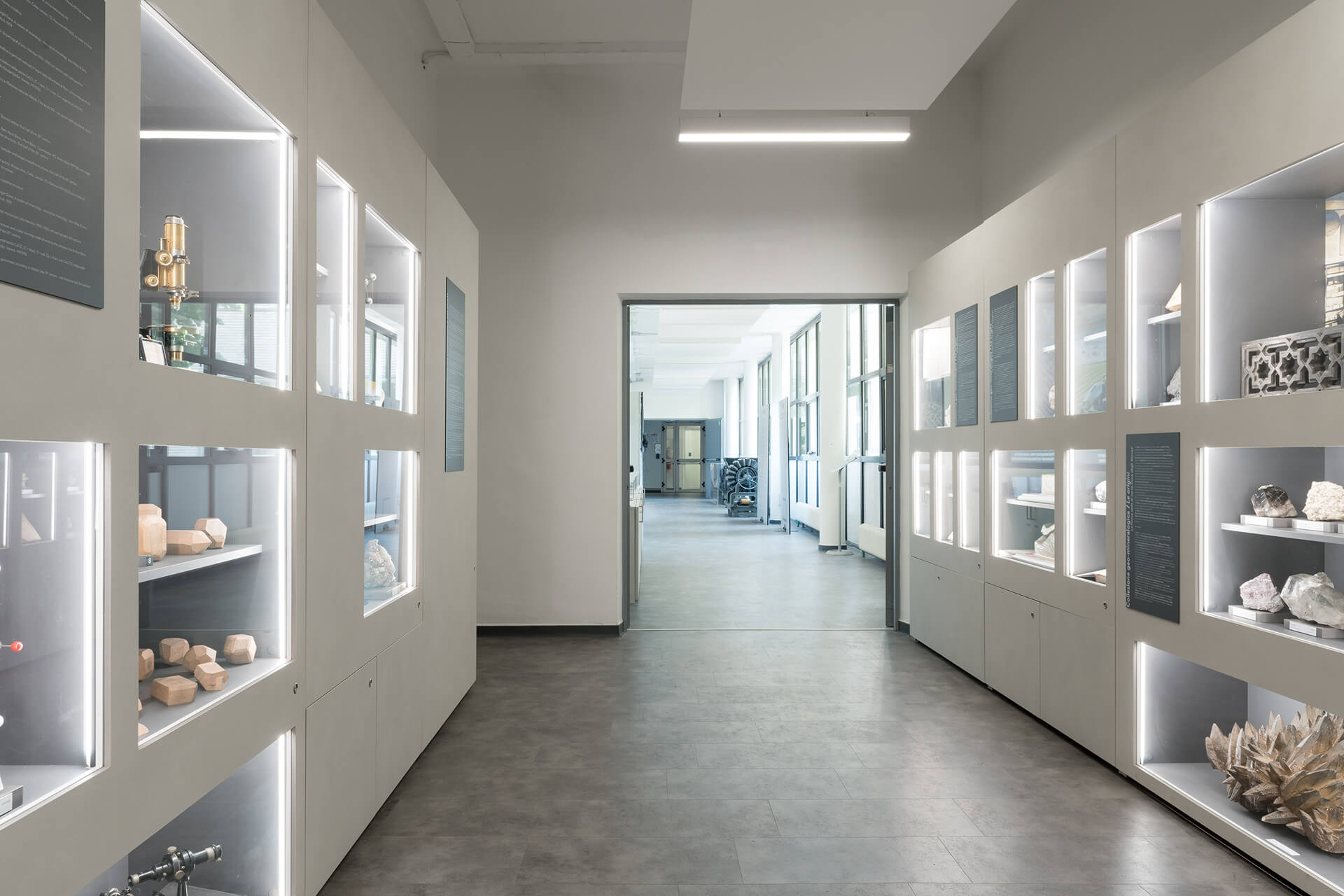 Vista interna di spazio espositivo organizzato come passaggio museale, caratterizzato da pareti attrezzate su entrambi i lati con moduli espositivi integrati in pannellature continue di colore chiaro. Le superfici sono scandite da nicchie illuminate incassate, contenenti campioni mineralogici, modelli geometrici, strumenti tecnici e reperti scientifici, disposti su supporti con etichette descrittive secondo un percorso tematico didattico. Accanto alle teche sono installati pannelli informativi verticali con testi di approfondimento, inseriti in un sistema grafico coordinato. L’illuminazione delle vetrine è integrata nella struttura dell’allestimento mediante profili luminosi lineari, mentre il soffitto presenta controsoffitti ribassati che ospitano i corpi illuminanti principali e gli impianti. La pavimentazione continua in materiale resiliente di colore neutro contribuisce alla lettura uniforme dello spazio, che si apre sullo sfondo verso un ambiente più ampio in cui sono visibili ulteriori macchinari esposti. L’immagine documenta un allestimento museale permanente progettato per la divulgazione scientifica e tecnologica, in cui arredi, grafica e sistemi illuminotecnici sono integrati in un unico dispositivo espositivo coerente.