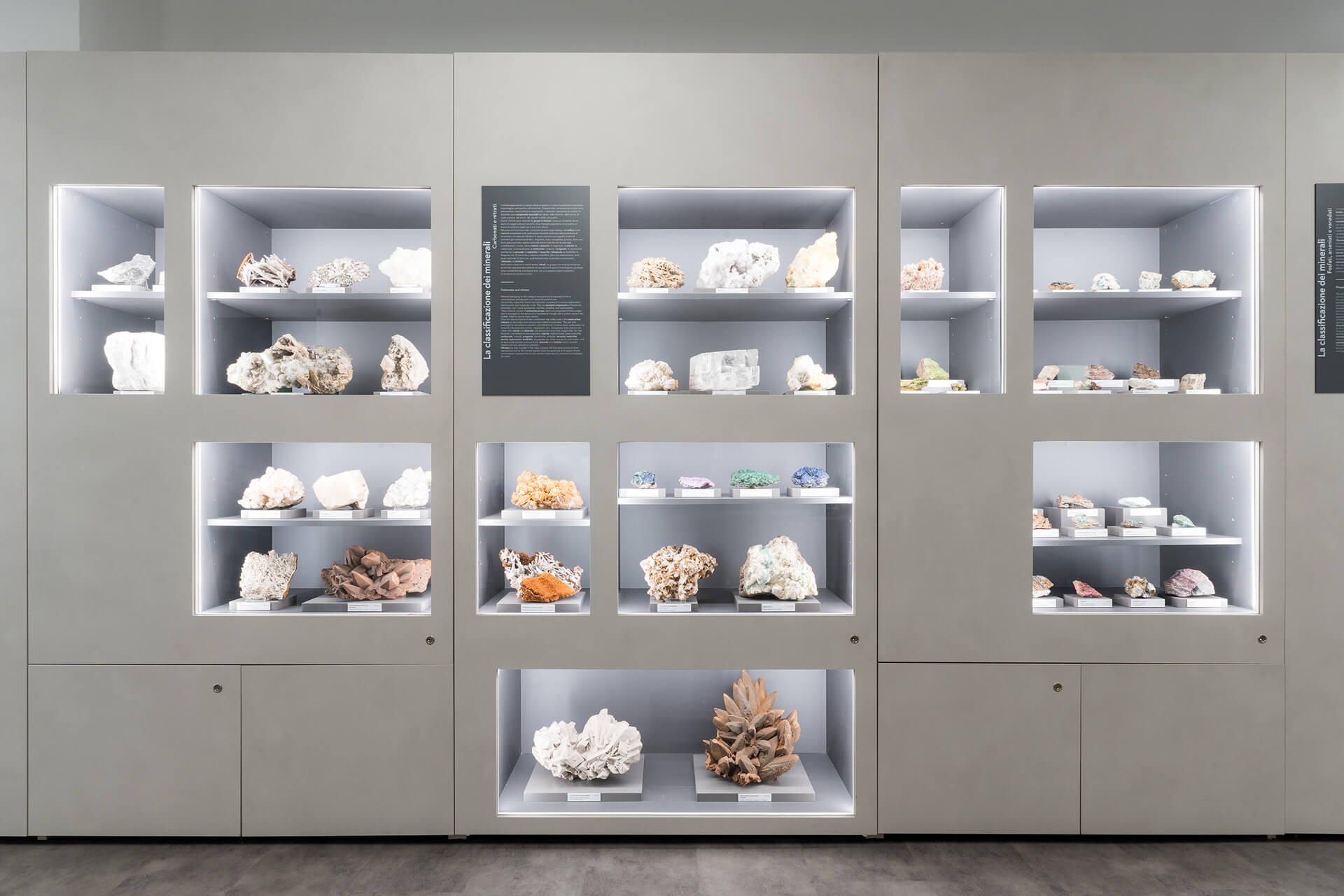 Vista frontale di modulo espositivo museale costituito da una parete attrezzata continua in pannellature di colore neutro, all’interno della quale sono ricavate nicchie illuminate incassate destinate all’esposizione di campioni mineralogici. Le teche, di diverse dimensioni e disposte secondo una griglia regolare, ospitano rocce e cristalli classificati, collocati su supporti con etichette identificative e organizzati per tipologia e caratteristiche mineralogiche. L’illuminazione è integrata nei vani espositivi mediante profili luminosi perimetrali che garantiscono una luce diffusa e uniforme, evidenziando texture, colore e morfologia dei campioni. Tra i moduli sono inseriti pannelli informativi verticali con testi esplicativi relativi alla classificazione dei minerali, integrati nel sistema grafico dell’allestimento. La parte inferiore della parete è costituita da sportelli tecnici complanari che nascondono vani di servizio e impianti, mentre la pavimentazione continua in materiale resiliente di tonalità neutra contribuisce alla lettura ordinata e didattica dello spazio. L’immagine documenta un allestimento museale permanente progettato per la divulgazione scientifica, in cui arredo, illuminazione e apparati informativi sono integrati in un unico sistema espositivo modulare.