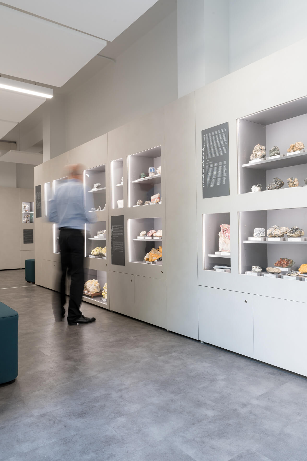 Vista interna di spazio espositivo museale con parete attrezzata continua destinata alla presentazione di campioni mineralogici, realizzata mediante pannellature modulari di colore chiaro integrate con nicchie illuminate incassate. All’interno delle teche sono esposti minerali e rocce classificati, disposti su supporti con etichette identificative e organizzati secondo criteri scientifici, accompagnati da pannelli informativi verticali con testi esplicativi relativi alla classificazione e alle proprietà dei materiali. L’illuminazione delle vetrine è ottenuta tramite profili luminosi lineari integrati nei bordi dei vani, mentre il soffitto presenta elementi ribassati e corpi illuminanti lineari sospesi che contribuiscono a una distribuzione uniforme della luce nello spazio. La pavimentazione continua in materiale resiliente di tonalità neutra definisce un ambiente ordinato e facilmente percorribile. Nell’immagine è visibile un visitatore in movimento davanti alle teche, elemento che evidenzia la fruizione dello spazio e la scala dell’allestimento. La configurazione complessiva documenta un allestimento museale permanente progettato per la divulgazione scientifica, in cui arredi espositivi, grafica informativa e impianti illuminotecnici sono integrati in un sistema unitario coerente.