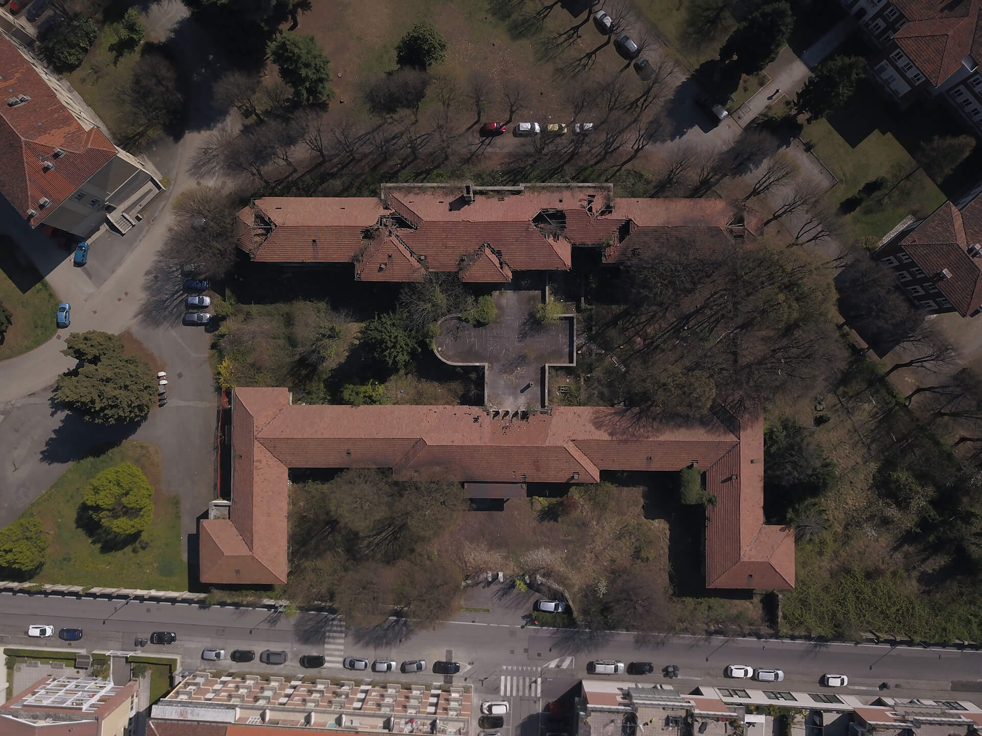 Questa immagine mostra una vista aerea di un complesso edilizio con un ampio cortile centrale, circondato da edifici con tetti a falde. La disposizione del complesso ricorda una forma a "C" con l'area centrale aperta. Il cortile sembra essere circondato da alberi e alcune aree verdi, mentre intorno sono visibili strade e veicoli parcheggiati. La parte inferiore dell'immagine mostra anche la zona residenziale circostante, con case o edifici in basso.