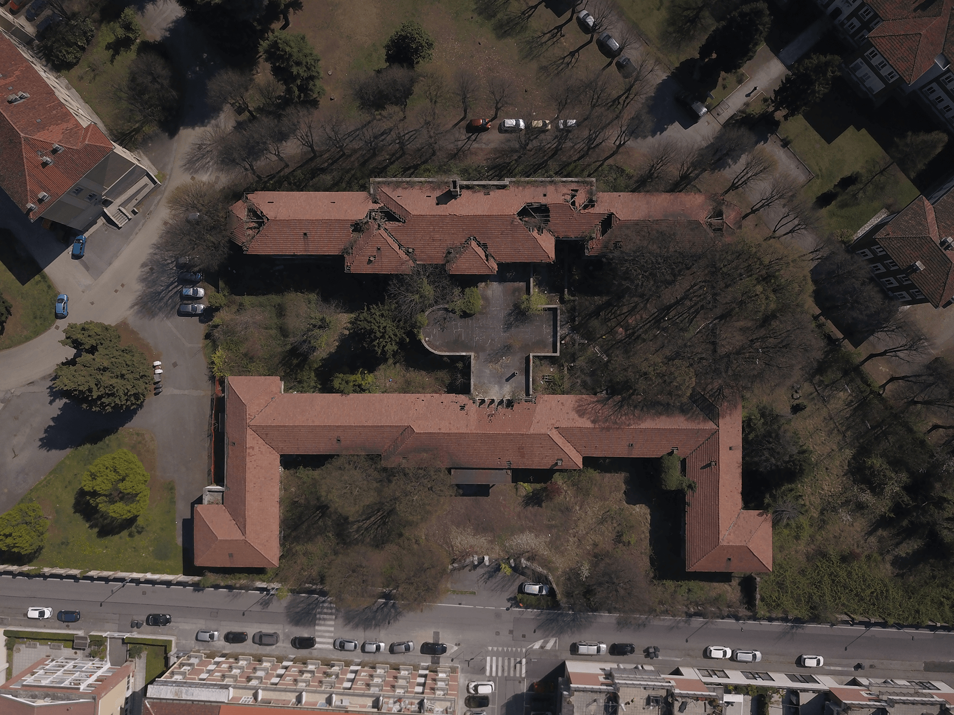 Questa immagine mostra una vista aerea di un complesso edilizio con un ampio cortile centrale, circondato da edifici con tetti a falde. La disposizione del complesso ricorda una forma a "C" con l'area centrale aperta. Il cortile sembra essere circondato da alberi e alcune aree verdi, mentre intorno sono visibili strade e veicoli parcheggiati. La parte inferiore dell'immagine mostra anche la zona residenziale circostante, con case o edifici in basso.