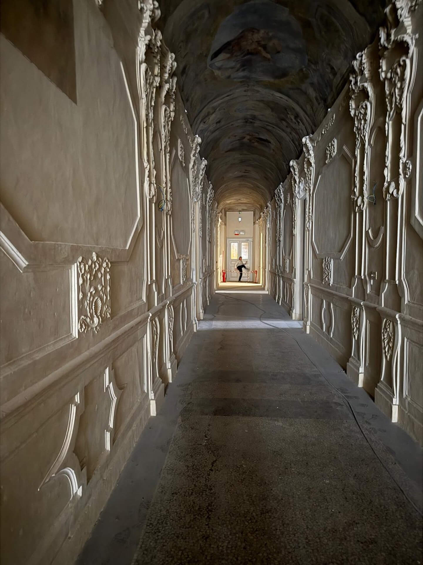 Corridoio interno di edificio storico con copertura a volta decorata e pareti laterali arricchite da stucchi ornamentali e cornici modanate di gusto barocco, caratterizzato da andamento longitudinale fortemente prospettico. Le superfici risultano in fase di restauro, con pavimentazione non finita e segni di intervento sulle pareti e sulla volta, mentre la luce naturale proveniente dall’apertura sul fondo evidenzia la ricchezza dei rilievi decorativi. Lo spazio documenta una fase di recupero conservativo dell’apparato architettonico originario, con impianti temporanei e cavi visibili lungo il percorso.