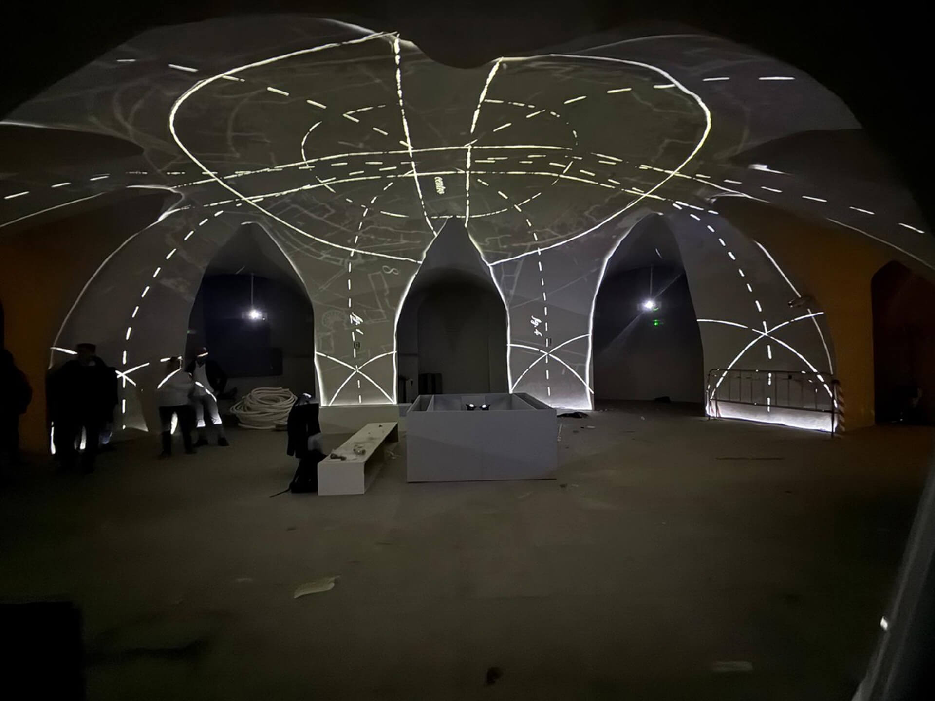 Ambiente interno voltato con copertura a crociere impostate su pilastri e archi in muratura, utilizzato come spazio espositivo all’interno di edificio storico riqualificato. Sulle superfici delle volte e delle pareti è proiettata una installazione luminosa con tracciati geometrici e linee di costruzione che seguono la geometria della struttura, evidenziando la forma delle campate e la scansione degli archi. Lo spazio, lasciato volutamente essenziale con pavimentazione continua e arredi minimi, mostra l’integrazione tra architettura storica restaurata e allestimento multimediale contemporaneo destinato a esposizione o installazione temporanea.