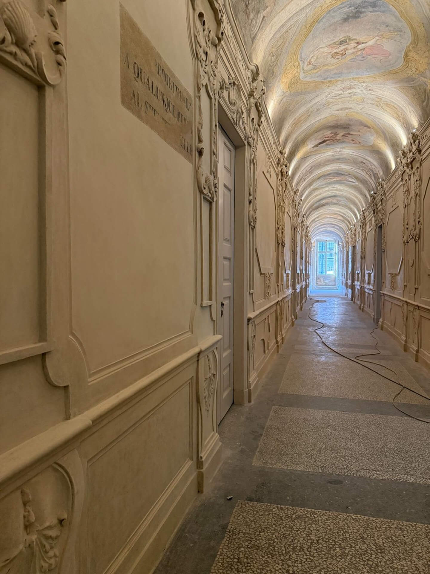 Corridoio monumentale di edificio storico con copertura a volta interamente affrescata e pareti decorate da stucchi ornamentali, cornici modanate e portali sagomati di gusto barocco, disposti in sequenza lungo l’asse longitudinale. Il pavimento in graniglia e le superfici restaurate indicano un intervento conservativo recente, mentre la luce naturale proveniente dalla finestra sul fondo enfatizza la profondità prospettica dello spazio e la ricchezza dell’apparato decorativo. La presenza di cavi temporanei lungo il pavimento suggerisce una fase di allestimento o completamento dei lavori all’interno del complesso storico.