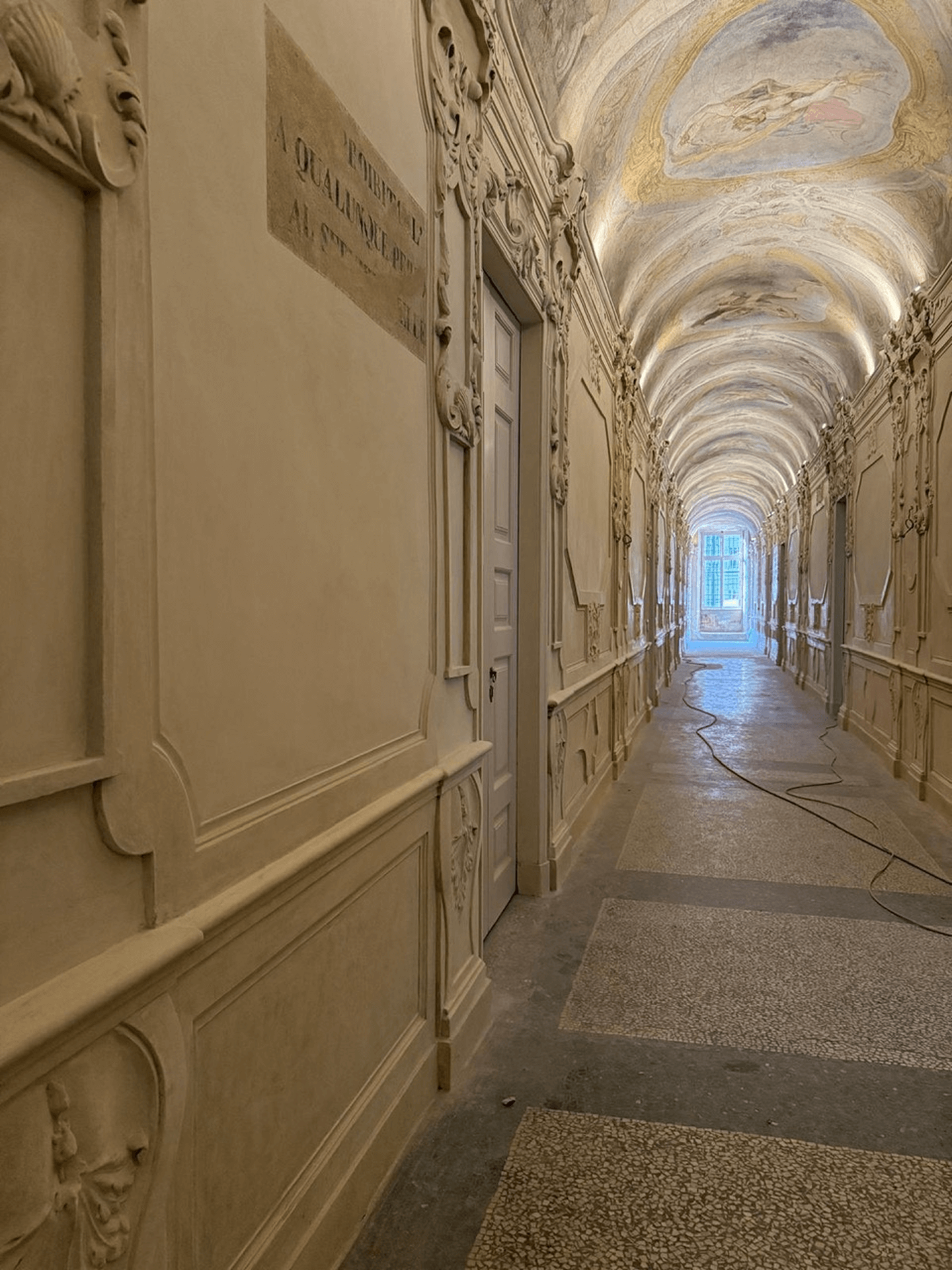 Corridoio monumentale con ricche decorazioni in stucco e soffitto affrescato restaurato, illuminato dalla luce proveniente da una finestra sul fondo.