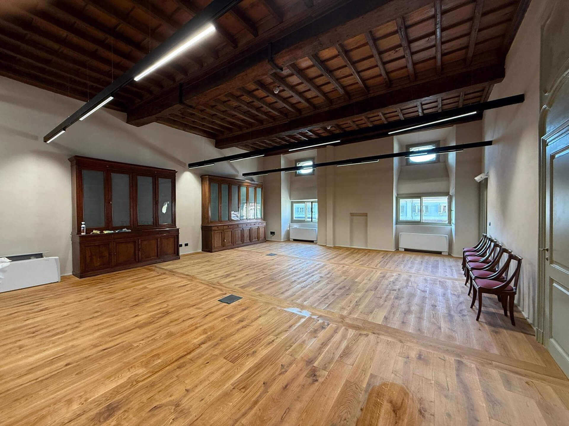 Sala interna di edificio storico restaurato, caratterizzata da soffitto ligneo con travi a vista e tavolato originale, pareti intonacate e pavimentazione in legno di nuova posa. Lungo le pareti sono presenti armadiature in legno integrate e aperture finestrate profonde che garantiscono illuminazione naturale, mentre corpi illuminanti lineari contemporanei sono sospesi sotto la struttura del solaio. Lo spazio, libero da arredi eccetto alcune sedute lungo il lato, mostra l’adattamento funzionale dell’ambiente a uso collettivo o didattico, evidenziando il dialogo tra elementi storici conservati e interventi impiantistici moderni.