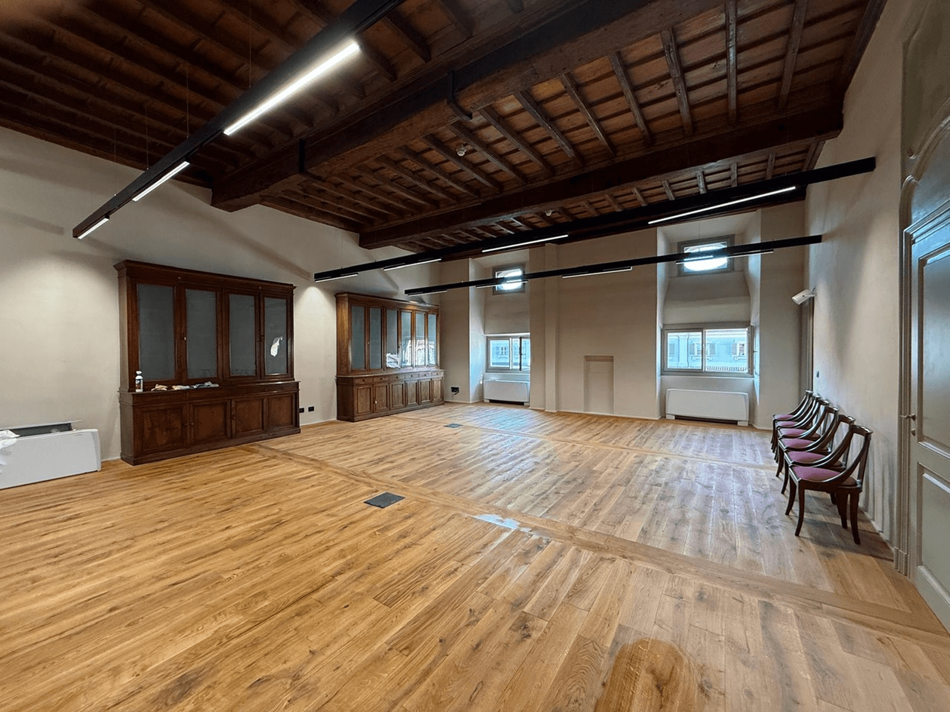 Ampia sala con pavimento in legno, soffitto con travi a vista e armadi storici lungo la parete, allestita per attività o incontri.