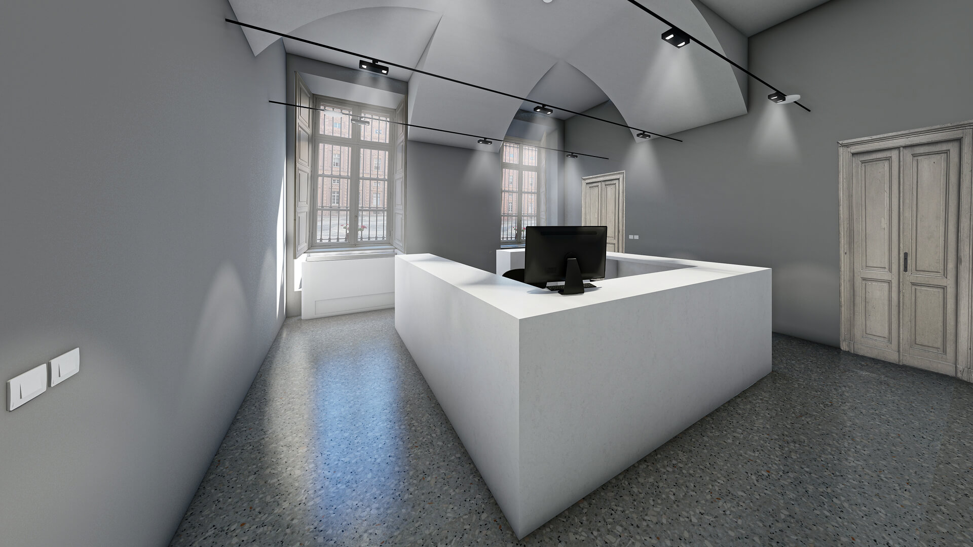 Ambiente interno restaurato con copertura a volte intonacate e pareti tinteggiate, caratterizzato da finestre profonde con infissi storici e pavimentazione in graniglia. Al centro dello spazio è inserito un volume contemporaneo monolitico di colore chiaro che funge da banco reception o postazione di lavoro, mentre l’illuminazione tecnica a binario è sospesa sotto le volte per valorizzare la geometria originaria del soffitto. La presenza di porte lignee storiche e finiture moderne evidenzia l’intervento di riqualificazione che integra nuovi elementi funzionali all’interno dell’architettura esistente.