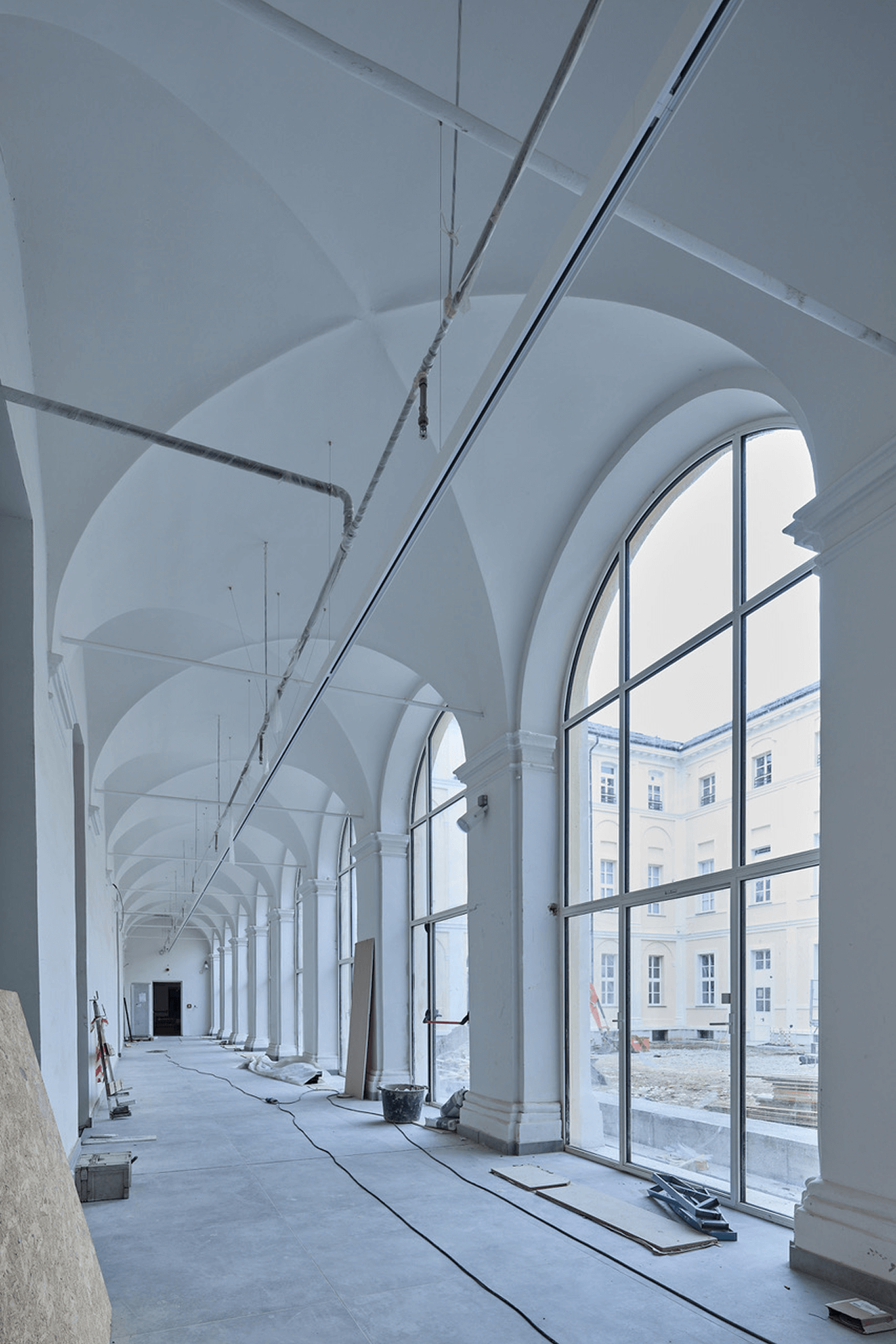 Corridoio porticato con volte a crociera e grandi finestre ad arco affacciate sul cortile, ripreso durante lavori di ristrutturazione con cavi e materiali di cantiere sul pavimento.