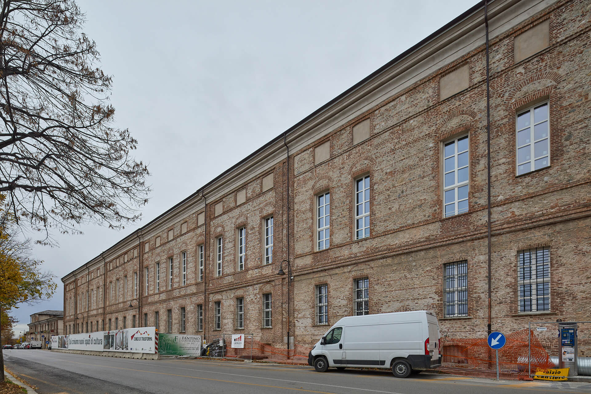 Prospetto longitudinale di edificio storico in muratura di laterizio a vista, caratterizzato da facciata modulare scandita da aperture rettangolari con cornici in mattoni e fasce marcapiano orizzontali. L’edificio si sviluppa su più livelli con copertura a falde e gronda aggettante, affacciato su strada urbana; lungo il fronte sono visibili recinzioni e segnaletica di cantiere che indicano un intervento di riqualificazione in corso, con presenza di mezzi e materiali temporanei lungo il marciapiede.