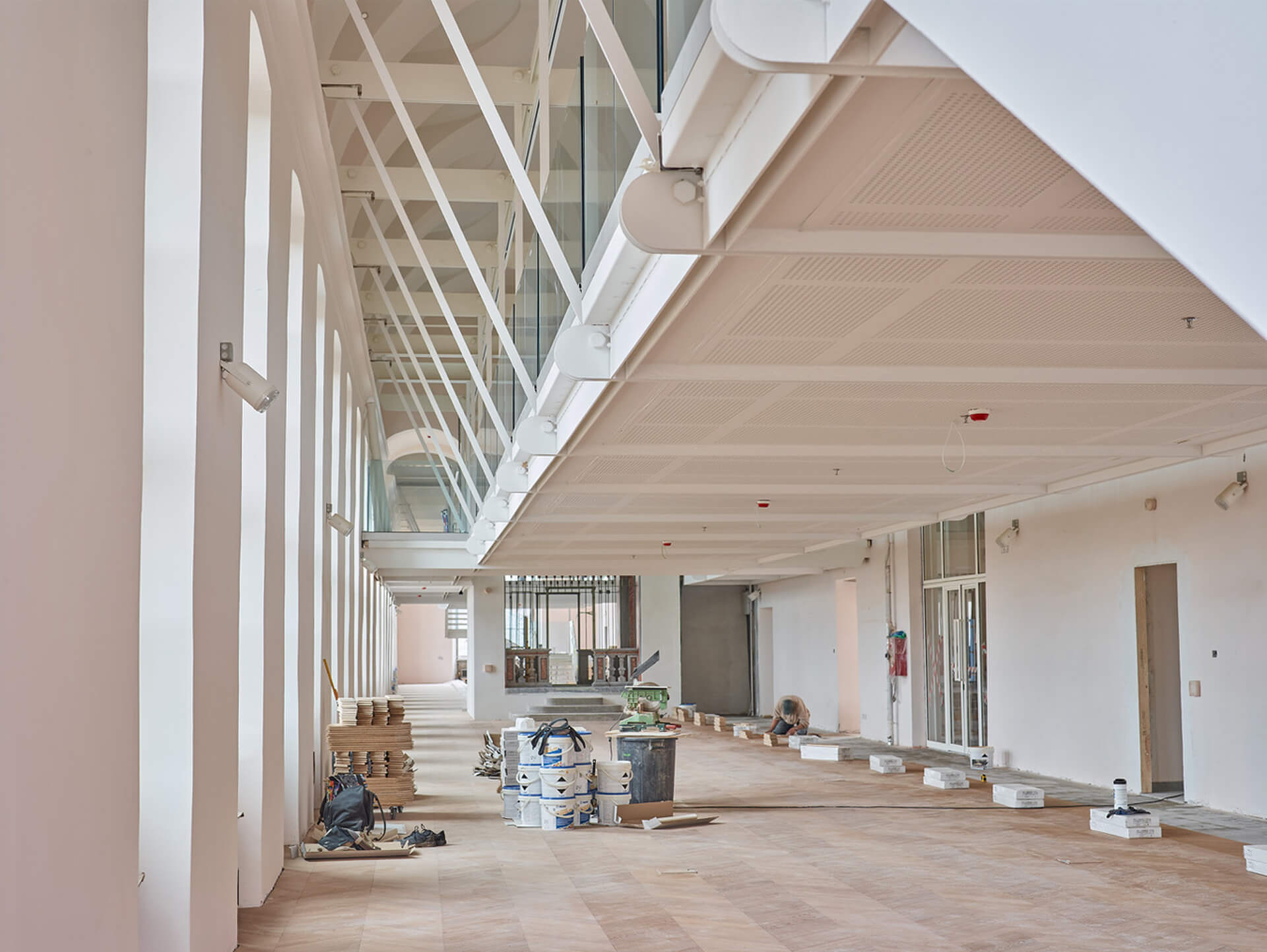 Spazio interno longitudinale oggetto di intervento di riqualificazione, caratterizzato da doppia altezza con struttura metallica verniciata di bianco che sostiene un ballatoio superiore con parapetti vetrati. Sul lato sinistro una sequenza di aperture ad arco scandisce il prospetto interno, mentre al piano terra sono visibili nuove pavimentazioni in posa, materiali da cantiere e attrezzature operative. L’ambiente presenta impianti tecnici integrati nella struttura del solaio e finiture in corso di completamento, evidenziando il dialogo tra elementi storici e inserimenti contemporanei.