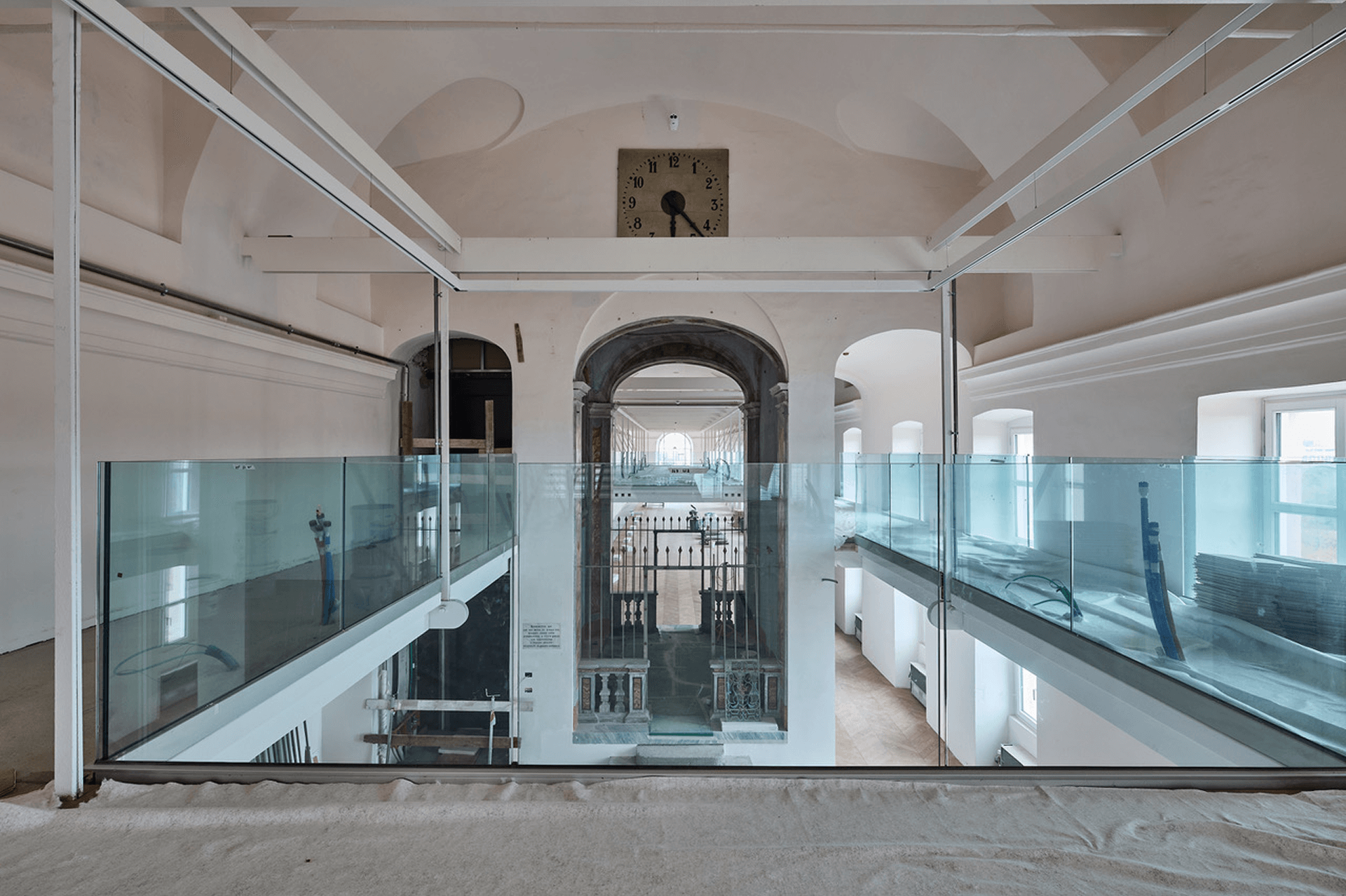 Scala interna con parapetti in vetro che collega due livelli di una grande sala con soffitto a volte e finestra ad arco, ripresa durante lavori di ristrutturazione.