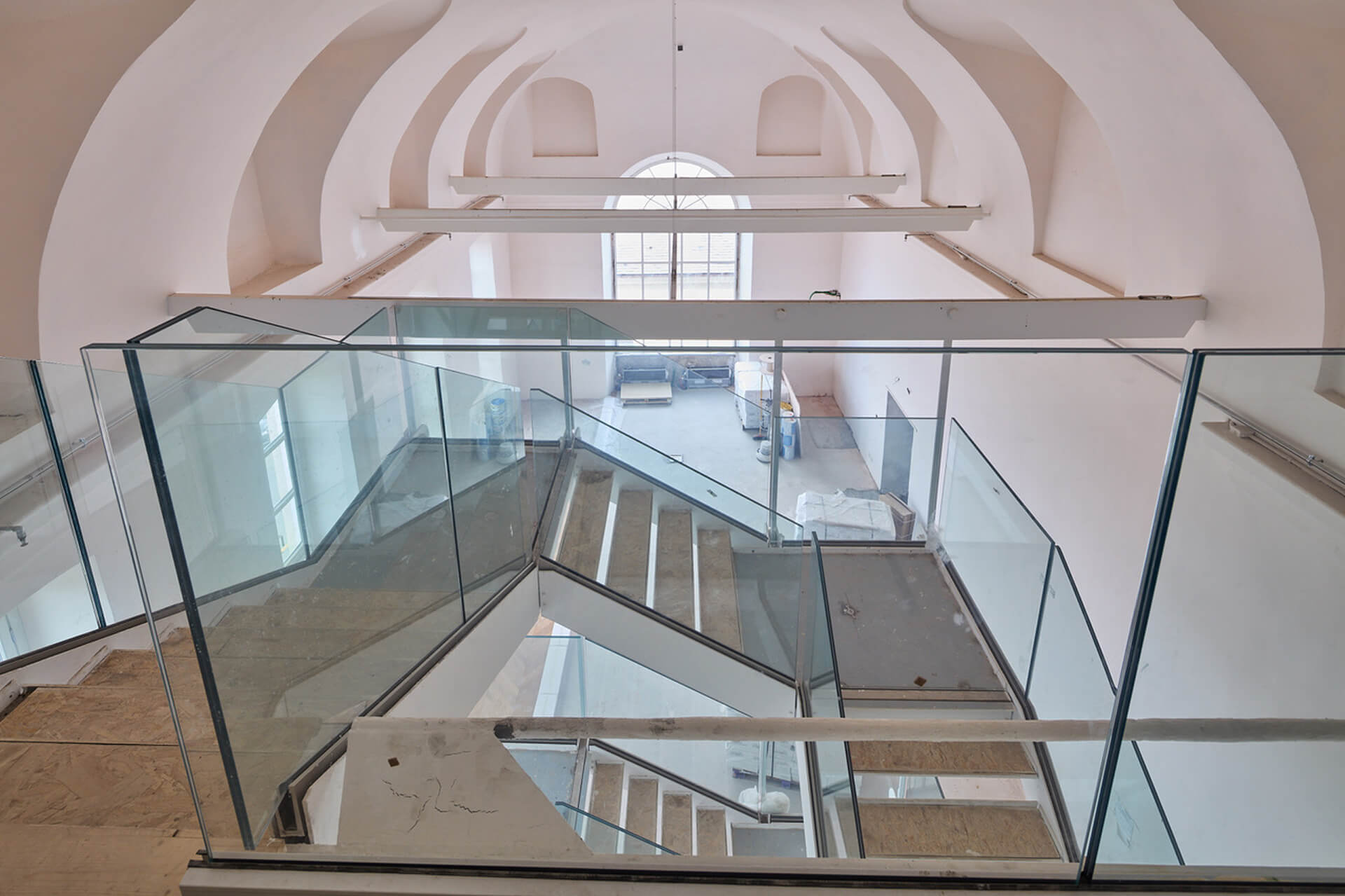 Ambiente interno voltato con copertura a botte intonacata, oggetto di intervento di riqualificazione con inserimento di nuovi collegamenti verticali in struttura metallica e parapetti in vetro. La scala contemporanea si sviluppa al centro dello spazio con rampe sovrapposte e pianerottoli sospesi, mentre le travi metalliche orizzontali collegano le pareti laterali senza alterare la geometria storica della volta. Sul fondo è visibile una grande apertura finestrata che illumina l’ambiente, evidenziando il contrasto tra le superfici originarie in muratura e i nuovi elementi architettonici in fase di completamento.