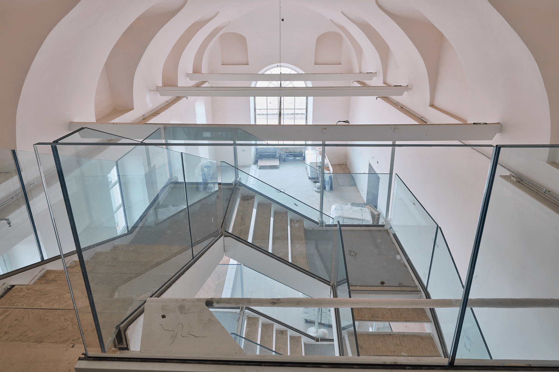 Scala interna con parapetti in vetro che collega due livelli di una grande sala con soffitto a volte e finestra ad arco, ripresa durante lavori di ristrutturazione.