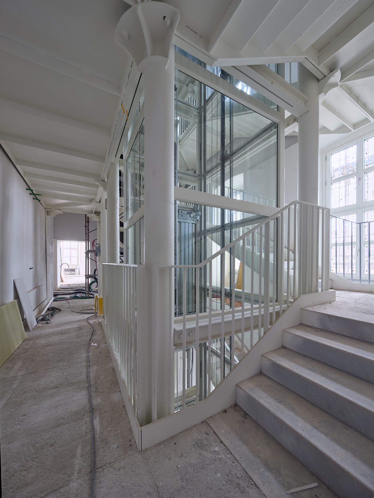 Spazio interno in fase di ristrutturazione caratterizzato da struttura portante con pilastri in ghisa e travi metalliche verniciate di bianco, all’interno del quale è stato inserito un nuovo vano ascensore con struttura in acciaio e tamponamenti vetrati. In primo piano una scala con parapetti metallici conduce al piano superiore, mentre lungo il corridoio sono visibili superfici non ancora finite, impianti temporanei e materiali di cantiere. Le grandi finestre laterali garantiscono illuminazione naturale, evidenziando il contrasto tra gli elementi strutturali storici e i nuovi interventi contemporanei in corso di completamento.