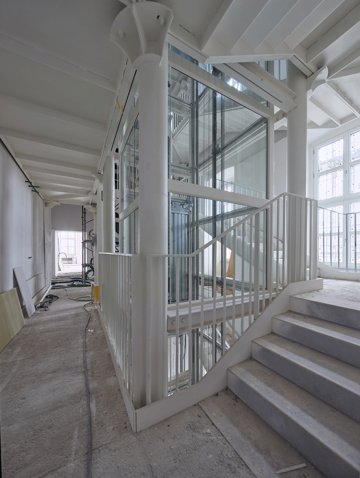 Scala interna accanto a un ascensore panoramico in vetro all’interno di un edificio in ristrutturazione, con struttura bianca, grandi finestre e attrezzature di cantiere sul pavimento.