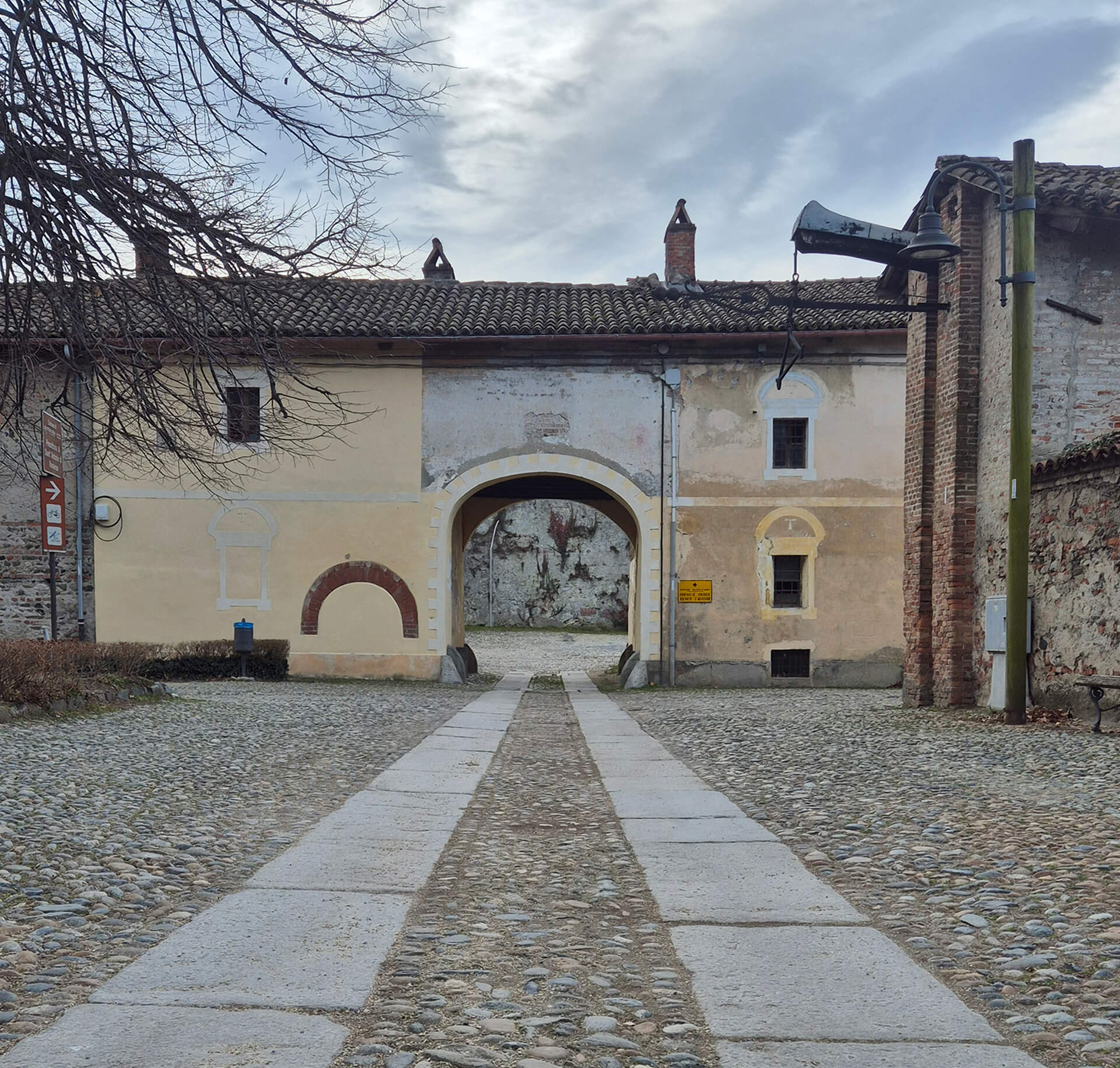 Fotografia di un edificio storico con accesso ad arco inserito in un contesto di corte pavimentata, caratterizzato da architettura tradizionale in muratura intonacata e copertura a falde con manto in coppi. Il prospetto principale è semplice e simmetrico, con un grande portale centrale ad arco ribassato che costituisce il passaggio verso uno spazio interno, probabilmente un cortile o un secondo recinto. La facciata presenta tracce di stratificazioni e interventi nel tempo, visibili nelle differenze di intonaco, nelle aperture tamponate e nelle cornici decorative parzialmente conservate. Ai lati del portale si riconoscono elementi architettonici dipinti o modellati nell’intonaco, come finte cornici e archi decorativi, che suggeriscono una configurazione originaria più articolata. Le piccole finestre con contorno sagomato e la presenza di camini sul tetto indicano un edificio di impianto antico, probabilmente legato a funzioni rurali, militari o di servizio. In primo piano è visibile una pavimentazione in ciottoli con percorso centrale in lastre di pietra, che conduce direttamente all’arco di ingresso e rafforza l’asse di simmetria della composizione. Ai lati sono presenti muri in pietra e mattoni, elementi metallici e corpi illuminanti, che testimoniano adattamenti contemporanei all’uso attuale dell’area. L’immagine documenta lo stato di conservazione dell’edificio e il rapporto tra architettura storica e spazio aperto, evidenziando la permanenza dei caratteri originari nonostante le trasformazioni e gli interventi successivi.
