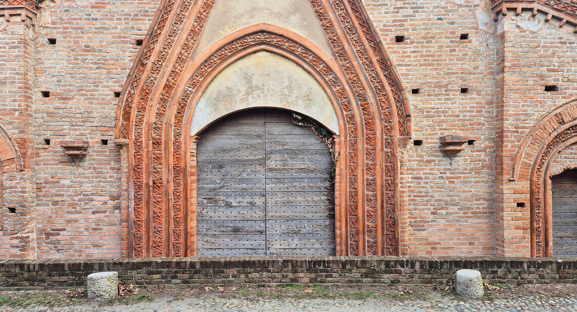 Dettaglio frontale della facciata in laterizio faccia a vista di un edificio storico di gusto neogotico, caratterizzata dalla presenza di aperture ad arco acuto con ricca decorazione in cotto modellato. L’immagine mostra un portale centrale con archivolto ogivale composto da più ghiere concentriche, realizzate con elementi sagomati e decorati con motivi vegetali e geometrici, che evidenziano una lavorazione artigianale di particolare pregio. Il portale è chiuso da un portone ligneo a due ante, costituito da tavole verticali rinforzate da chiodature metalliche, inserito all’interno di una cornice arretrata rispetto al filo della facciata. Sopra l’arco è presente una porzione intonacata, probabilmente frutto di un intervento successivo o di una perdita dell’originario rivestimento, che interrompe la continuità del paramento in laterizio. La muratura circostante è realizzata con mattoni pieni disposti in corsi regolari, con presenza di piccoli fori e alloggiamenti che potrebbero essere legati a ponteggi originari o a elementi costruttivi rimossi. Ai lati del portale si notano mensole in laterizio sporgenti e modanature decorative che contribuiscono alla composizione simmetrica della facciata. In primo piano è visibile un basso muretto in laterizio che separa l’edificio dallo spazio esterno pavimentato in ciottoli, mentre la superficie muraria mostra segni di alterazione cromatica, erosione dei giunti e depositi superficiali, compatibili con l’esposizione prolungata agli agenti atmosferici. L’immagine documenta con chiarezza le caratteristiche stilistiche, la qualità costruttiva e lo stato di conservazione dell’apparato decorativo in cotto, elementi di particolare interesse per l’analisi storico-architettonica e per eventuali interventi di restauro conservativo.