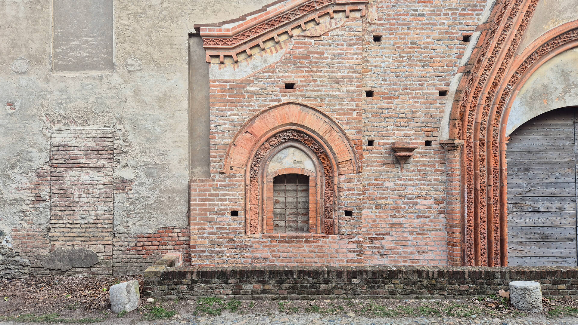 Dettaglio della facciata in laterizio decorato di un edificio storico con caratteri neogotici, in cui si evidenzia chiaramente la sovrapposizione di fasi costruttive differenti e il rapporto tra una struttura più antica intonacata e un successivo intervento in mattoni faccia a vista di maggiore qualità architettonica. Al centro dell’immagine è visibile una piccola apertura ad arco acuto, incorniciata da più ghiere in cotto sagomato con motivi ornamentali vegetali e geometrici, realizzate con elementi modellati di notevole precisione. L’arco è inserito all’interno di un riquadro murario in laterizio disposto in corsi regolari, che contrasta con la parete adiacente sulla sinistra, intonacata e fortemente degradata, dove sono evidenti tracce di tamponamenti e rifacimenti successivi, riconoscibili dalla diversa tessitura e dal colore dei materiali. Sopra l’apertura si sviluppa una cornice decorativa in cotto con motivi dentellati e modanature, che segue l’andamento inclinato della copertura e sottolinea la composizione architettonica della facciata. La qualità della lavorazione e la ricchezza dell’apparato ornamentale indicano un intervento di carattere rappresentativo, probabilmente successivo rispetto alla muratura più povera visibile a sinistra. Sulla destra compare parte del portale principale, anch’esso ad arco ogivale con ghiera decorata, chiuso da un portone ligneo, che conferma l’unitarietà stilistica dell’intervento in laterizio. In primo piano è presente un basso muretto di contenimento in mattoni che separa la facciata dallo spazio esterno pavimentato in ciottoli, mentre alla base delle murature si notano fenomeni di alterazione cromatica, erosione dei giunti e depositi superficiali dovuti all’umidità e all’esposizione agli agenti atmosferici. L’immagine consente una chiara lettura delle stratificazioni costruttive, delle differenze materiche e dello stato di conservazione, risultando particolarmente significativa per analisi storico-architettoniche e per la definizione di interventi di restauro conservativo.