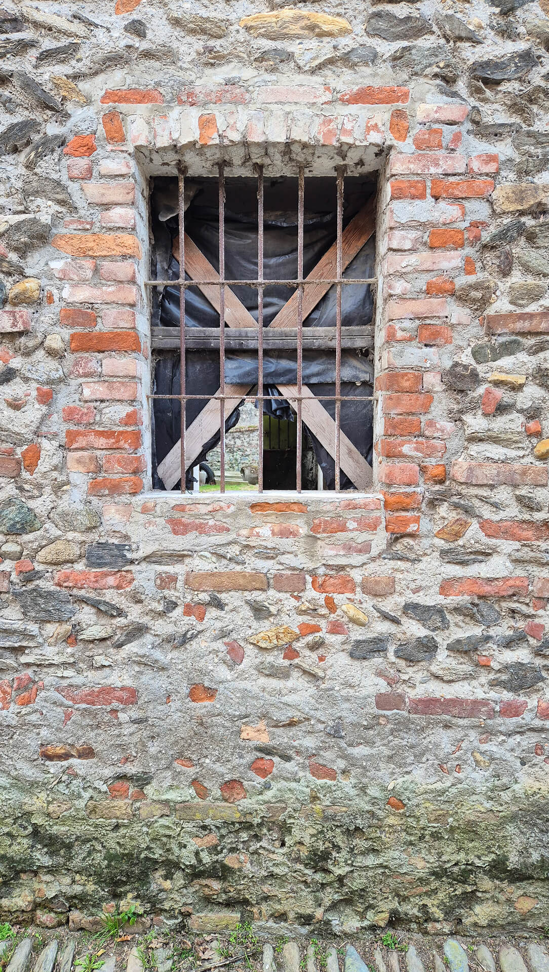 Dettaglio ravvicinato di una apertura finestrata inserita in muratura mista storica, costituita da pietrame irregolare, ciottoli e frammenti di laterizio legati con malta di calce, con evidenti segni di degrado superficiale e di interventi successivi. Il vano presenta una cornice in mattoni pieni disposti con maggiore regolarità rispetto al paramento circostante, a indicare una fase costruttiva differente o un rifacimento dell’apertura rispetto alla muratura originaria. La parte superiore della finestra è chiusa da un arco ribassato in laterizio, realizzato con elementi disposti radialmente, mentre lateralmente i piedritti mostrano integrazioni e sostituzioni di mattoni, riconoscibili per differenze cromatiche e di tessitura. All’interno del vano è presente una grata metallica verticale, fortemente ossidata, che testimonia la funzione di sicurezza o aerazione dell’ambiente retrostante. Dietro l’inferriata si notano elementi lignei disposti a croce e un telo di protezione, segni di una chiusura provvisoria o di uno stato di abbandono. La superficie muraria presenta diffusi fenomeni di alterazione, tra cui erosione dei giunti, perdita di malta, disgregazione dei materiali e presenza di umidità nella fascia basamentale, dove si osservano incrostazioni, colonizzazioni biologiche e depositi dovuti al contatto diretto con il terreno. Alla base della parete è visibile una pavimentazione esterna in ciottoli, tipica degli spazi di corte, che contribuisce al ristagno dell’acqua e all’aggravarsi del degrado. L’immagine è particolarmente significativa per la lettura della tecnica costruttiva tradizionale, delle stratificazioni murarie e dello stato di conservazione, fornendo indicazioni utili per analisi diagnostiche, rilievo materico e valutazioni preliminari in vista di interventi di restauro o consolidamento.