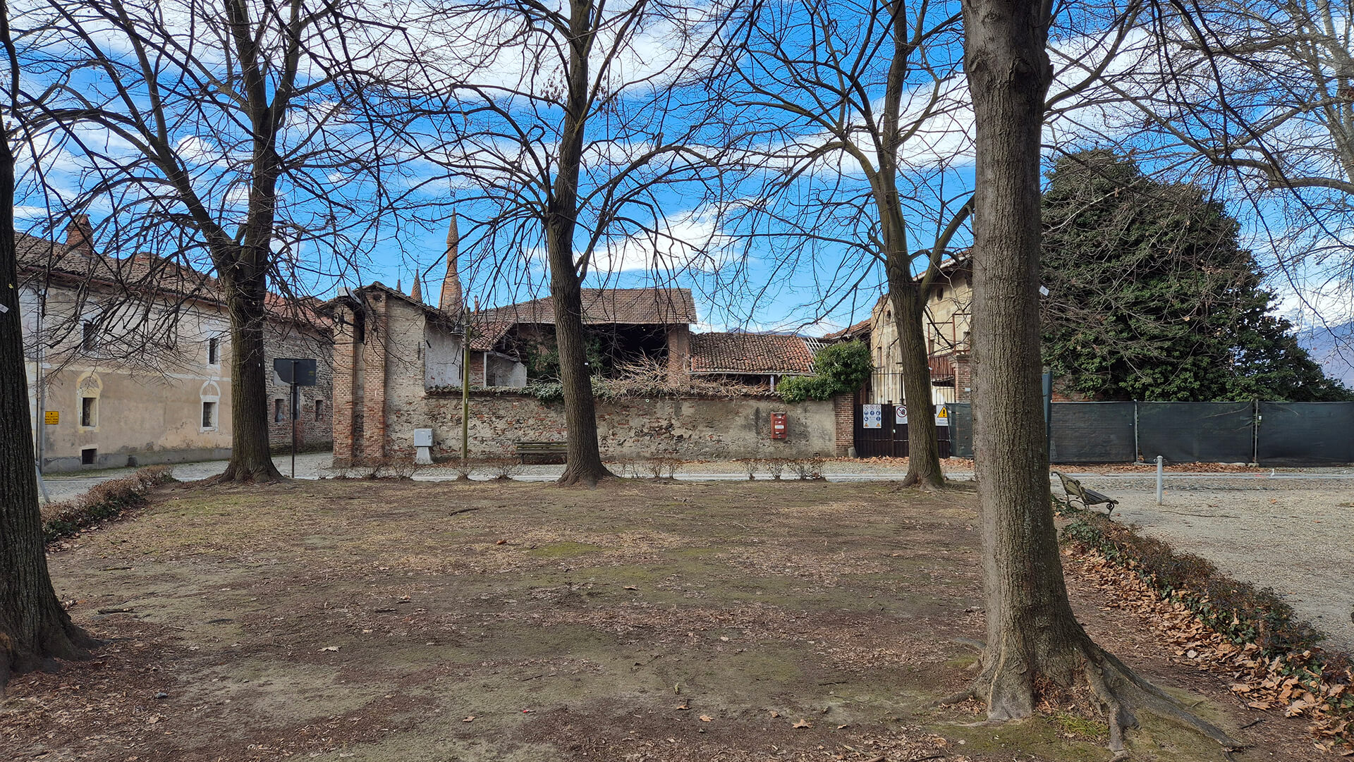 Vista d’insieme di uno spazio aperto alberato situato di fronte a un complesso edilizio storico di impianto rurale, delimitato da un muro di cinta in muratura mista e da edifici con copertura a falde in coppi. In primo piano si sviluppa un’area verde pianeggiante, priva di pavimentazione, caratterizzata da terreno battuto con presenza di foglie secche e radici affioranti, occupata da alberi ad alto fusto disposti in filare, che definiscono uno spazio di pertinenza esterno probabilmente utilizzato come area di sosta o zona filtro tra la strada e il complesso. Sullo sfondo è visibile il muro perimetrale in pietrame e laterizio, con andamento continuo e altezza contenuta, che separa lo spazio pubblico dalla corte interna. Al centro del muro si riconosce un accesso carrabile con cancello metallico, mentre lungo la recinzione sono presenti elementi funzionali come cassette postali, segnaletica e pannelli informativi, che indicano un uso attuale parziale o controllato dell’area. Oltre il muro emergono i corpi di fabbrica del complesso, costituiti da edifici di diversa epoca e qualità costruttiva, con murature intonacate o in laterizio a vista, porticati e coperture tradizionali in tegole. Tra le coperture si intravedono le torrette in laterizio di carattere decorativo, già osservate in altri dettagli, che contribuiscono a identificare l’edificio di maggiore pregio architettonico all’interno del complesso. La presenza di alberature mature, arredi urbani e superfici non pavimentate suggerisce che lo spazio antistante abbia assunto nel tempo una funzione di piazzale o piccolo giardino pubblico, pur mantenendo il rapporto diretto con l’impianto storico originario. L’immagine risulta utile per la lettura del rapporto tra spazio aperto, muro di cinta e complesso edilizio, evidenziando il contesto ambientale, lo stato di conservazione generale e le relazioni tra area pubblica e struttura storica, elementi fondamentali per analisi urbanistiche, rilievo architettonico e ipotesi di recupero o rifunzionalizzazione.