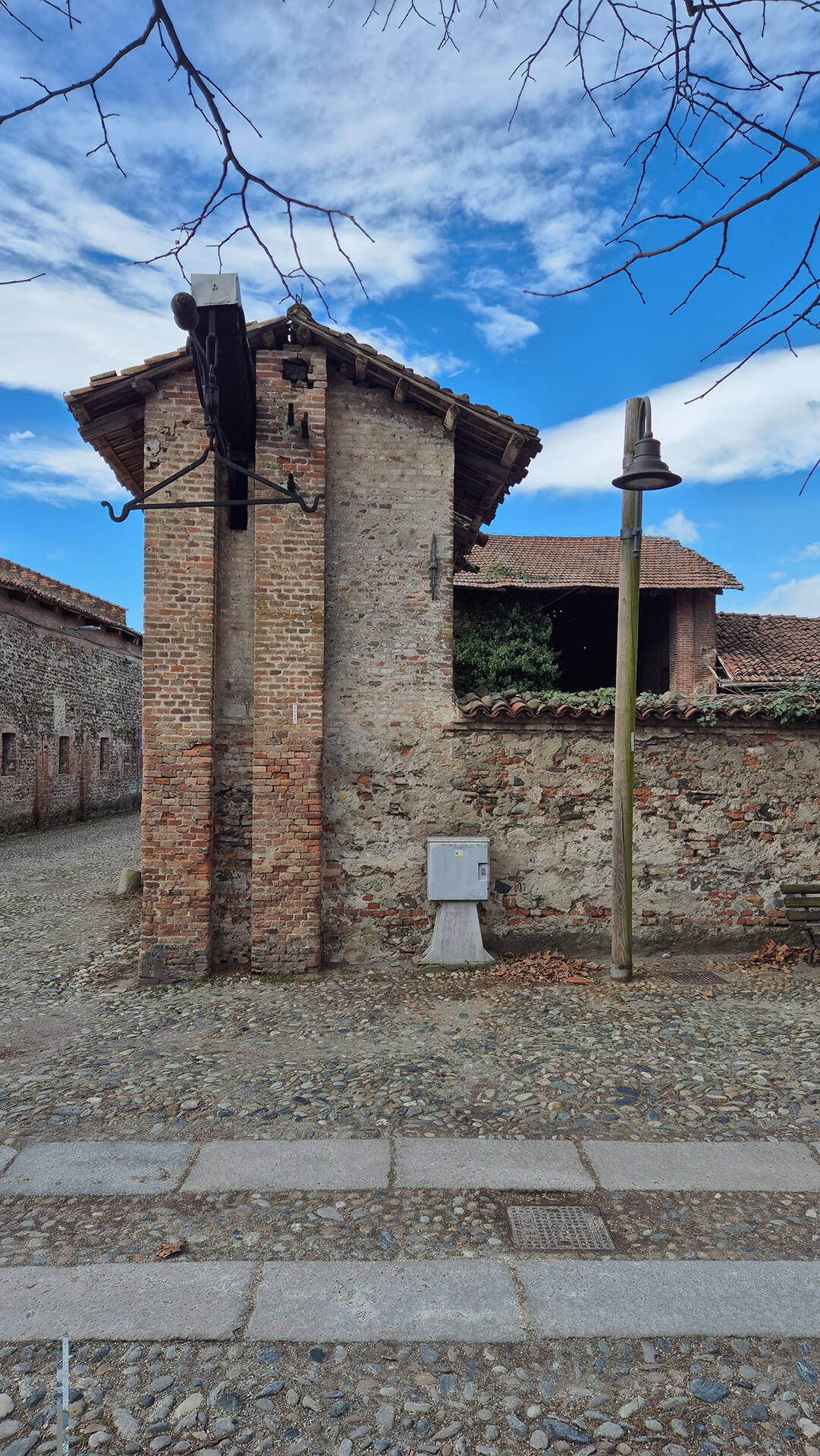 Vista frontale di un elemento architettonico in muratura appartenente al sistema di accesso o di servizio del complesso storico, costituito da un corpo verticale in laterizio pieno addossato al muro di cinta perimetrale realizzato in muratura mista di pietrame e mattoni legati con malta. Il volume presenta andamento stretto e sviluppato in altezza, concluso superiormente da una piccola copertura a falde con struttura lignea e manto in coppi, in continuità con le coperture tradizionali degli edifici circostanti. Sulla facciata è presente una struttura metallica sporgente fissata alla muratura, composta da staffe e braccio orizzontale, probabilmente residuo di un antico sistema di illuminazione, segnalazione o supporto funzionale legato all’accesso al complesso. La tessitura muraria evidenzia chiaramente interventi stratificati, con porzioni in laterizio regolare alternate a tratti in muratura irregolare, indicativi di modifiche e riparazioni successive. Il muro di cinta retrostante, intonacato in parte e in parte lasciato a vista, delimita la corte interna, dalla quale emergono coperture in coppi e volumi rurali o di servizio, riconducibili all’impianto originario del complesso. In primo piano la pavimentazione è realizzata in acciottolato con inserti in lastre lapidee, tipica delle sistemazioni storiche di corti e spazi di pertinenza, mentre sulla destra è visibile un palo di illuminazione contemporaneo e una panchina, elementi che testimoniano l’attuale uso pubblico o semi-pubblico dell’area. L’immagine consente di leggere con chiarezza il rapporto tra muro perimetrale, elementi di accesso e strutture di servizio, mettendo in evidenza lo stato di conservazione delle murature storiche, la presenza di aggiunte moderne e la continuità materica tra i diversi edifici del complesso, aspetti rilevanti per analisi architettonica, documentazione dello stato di fatto e valutazioni di restauro o recupero funzionale.