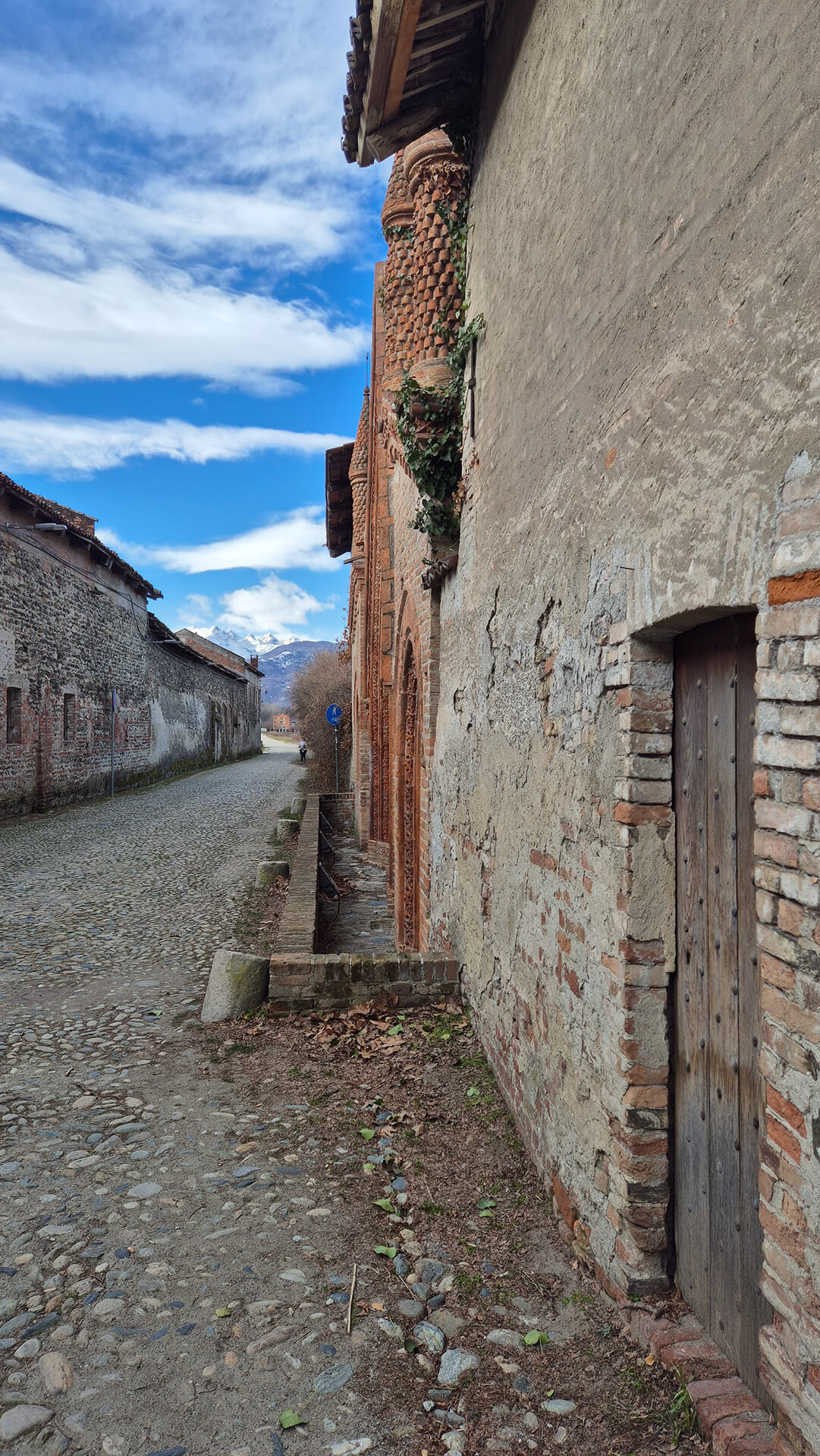 Vista prospettica lungo un tracciato viario storico pavimentato in acciottolato, affiancato da edifici appartenenti al complesso edilizio di impianto rurale e religioso già documentato nelle immagini precedenti. In primo piano, sulla destra, è visibile una muratura intonacata con evidenti fenomeni di degrado superficiale, lacune dell’intonaco e porzioni di laterizio a vista, nella quale si apre una porta secondaria con architrave in laterizio e battente ligneo, probabilmente accesso di servizio a locali interni. Procedendo lungo la parete si riconosce il prospetto laterale dell’edificio in laterizio decorato, caratterizzato da lesene, cornici e torrette cilindriche in mattoni sagomati, elementi ornamentali di gusto neogotico già osservati in altre viste. Alla base della facciata corre un piccolo canale di scolo in muratura, destinato allo smaltimento delle acque meteoriche, che separa l’edificio dalla strada e testimonia l’impianto originario di tipo rurale o conventuale. Sul lato opposto della strada si sviluppa un lungo edificio in muratura mista di pietrame e laterizio, con piccole aperture regolari, probabilmente destinato in origine a funzioni agricole o di deposito, che contribuisce a definire un ambito viario stretto e lineare di carattere storico, conservato nella pavimentazione tradizionale e nell’assenza di sistemazioni moderne invasive. Sullo sfondo si intravede il paesaggio collinare e montano, che colloca il complesso in un contesto territoriale periferico o di margine urbano, mentre la presenza di segnaletica e di percorsi ciclopedonali indica un uso attuale dell’area compatibile con la fruizione pubblica. L’immagine risulta significativa per la lettura del rapporto tra edificio decorato, muro di cinta, viabilità storica e contesto paesaggistico, mettendo in evidenza la stratificazione costruttiva, lo stato di conservazione delle superfici murarie e la permanenza dell’impianto originario, elementi utili per analisi architettonica, rilievo dello stato di fatto e valutazioni di restauro o riqualificazione.