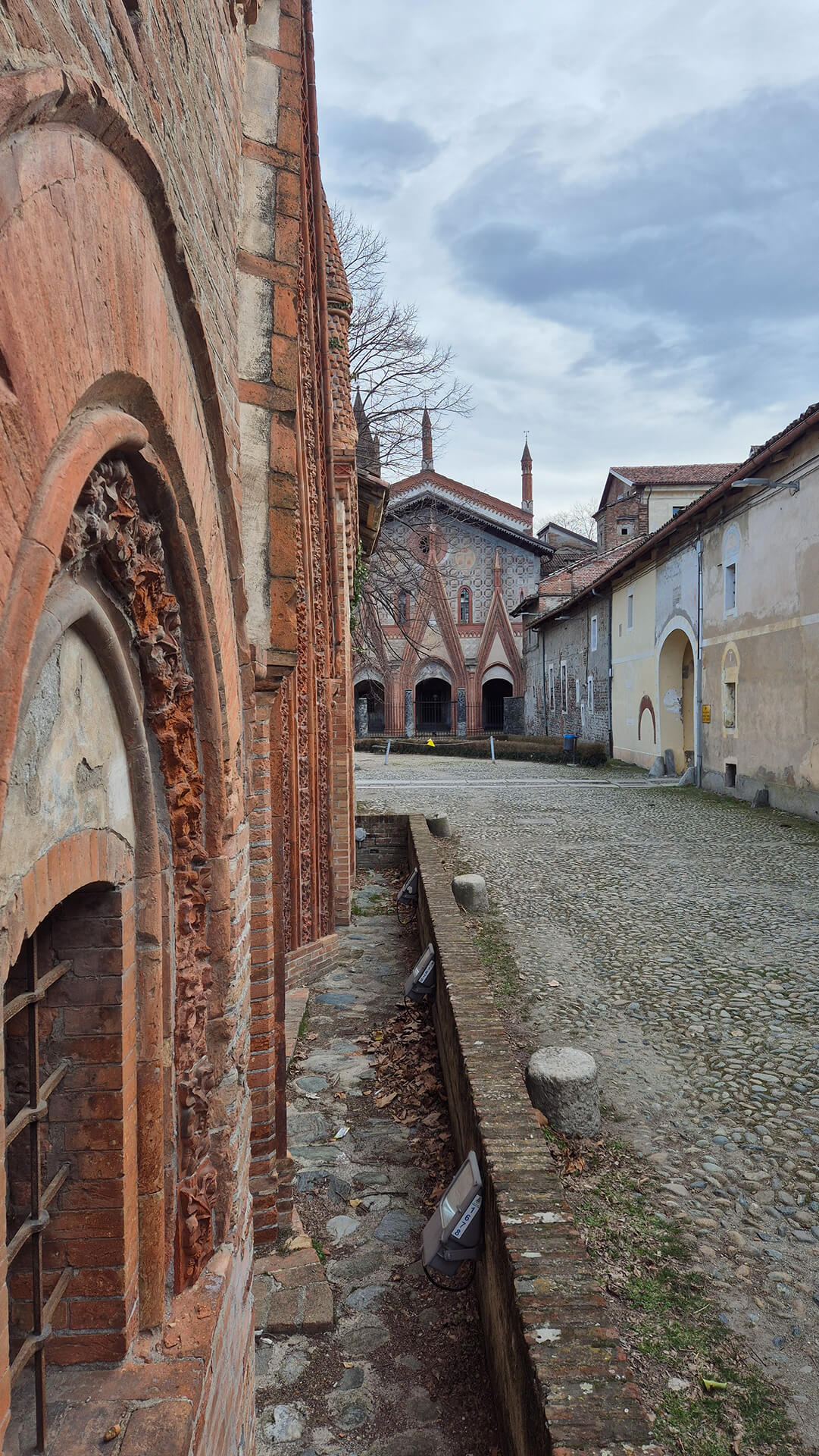 Vista prospettica lungo il prospetto laterale dell’edificio in laterizio decorato, con inquadratura radente che mette in evidenza la successione delle aperture ad arco ogivale strombato e la ricchezza dell’apparato ornamentale in cotto, costituito da ghiere multiple modanate e fasce decorate con motivi fitomorfi a rilievo. La muratura è realizzata in laterizio faccia a vista con corsi regolari, interrotta da elementi sagomati e lesene che scandiscono verticalmente il prospetto, confermando il carattere neogotico dell’edificio. Alla base della facciata corre un canale di drenaggio in muratura, leggermente ribassato rispetto al piano della corte, destinato allo smaltimento delle acque meteoriche; tale elemento separa il fabbricato dalla pavimentazione esterna in acciottolato e testimonia l’impostazione originaria dell’area come corte rurale o conventuale. Sono visibili anche apparecchi illuminanti contemporanei appoggiati lungo il bordo del canale, indicativi di un uso attuale dello spazio per attività di visita o valorizzazione. L’inquadratura si apre verso la corte interna pavimentata in ciottoli, delimitata da edifici di diversa epoca e tipologia costruttiva: sulla destra si riconoscono corpi di fabbrica intonacati con aperture ad arco ribassato, mentre sullo sfondo emerge il prospetto principale dell’edificio monumentale, caratterizzato da decorazioni in laterizio, porticati ad archi acuti e torrette slanciate, elementi già osservati nei dettagli precedenti. La presenza di superfici murarie con differenti finiture — laterizio a vista, intonaco degradato, inserti lapidei — evidenzia la stratificazione storica del complesso, probabilmente costituito da un nucleo originario di pregio architettonico affiancato da edifici di servizio realizzati in epoche successive. L’immagine risulta particolarmente utile per la lettura del rapporto tra facciata decorata, corte interna e corpi edilizi secondari, mettendo in evidenza la configurazione spaziale del complesso, le tecniche costruttive tradizionali e lo stato di conservazione delle superfici, elementi fondamentali per analisi storico-architettoniche, rilievo dello stato di fatto e valutazioni di restauro o rifunzionalizzazione.