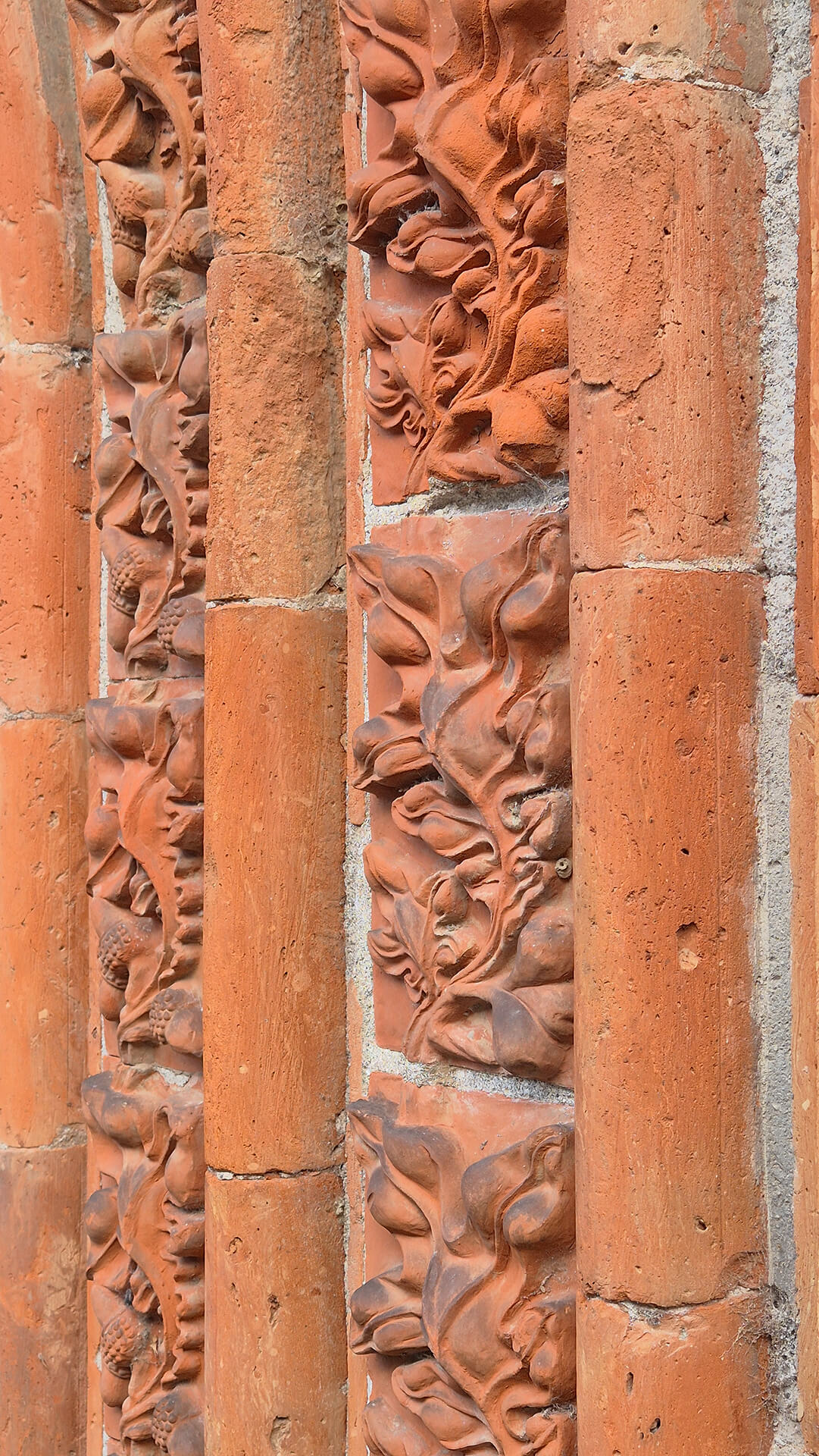 Dettaglio ravvicinato dell’apparato decorativo in laterizio sagomato appartenente alla strombatura di un portale o di una finestra ogivale dell’edificio di carattere neogotico, nel quale sono visibili le fasce ornamentali costituite da elementi in cotto modellati a rilievo con motivi fitomorfi. I singoli moduli decorativi raffigurano foglie, tralci e frutti stilizzati disposti in sequenza verticale, inseriti tra sottili colonnine cilindriche anch’esse realizzate in laterizio, che scandiscono ritmicamente la superficie e contribuiscono alla definizione plastica dell’archivolto. Le colonnine, composte da conci cilindrici sovrapposti, presentano superfici leggermente irregolari e porose, con variazioni cromatiche dal rosso aranciato al bruno, dovute sia alla cottura del materiale sia ai fenomeni di ossidazione e deposito superficiale. Tra gli elementi decorativi sono visibili le fughe di malta, in parte erose, che rendono leggibile la tecnica costruttiva basata sull’assemblaggio di pezzi prefabbricati in cotto successivamente messi in opera. Lo stato di conservazione mostra abrasioni, piccole lacune e arrotondamenti dei rilievi, soprattutto nelle parti più sporgenti, oltre a microfessurazioni e discontinuità nelle giunzioni, compatibili con l’esposizione agli agenti atmosferici e con l’età del manufatto. Nonostante ciò, la qualità della modellazione rimane chiaramente percepibile, evidenziando una lavorazione artigianale di notevole precisione. L’immagine è particolarmente significativa per lo studio del dettaglio ornamentale in cotto, delle tecniche di prefabbricazione e della composizione modulare dell’apparato decorativo, fornendo informazioni utili per analisi storico-architettoniche, rilievo materico e valutazioni di restauro conservativo dell’edificio. 