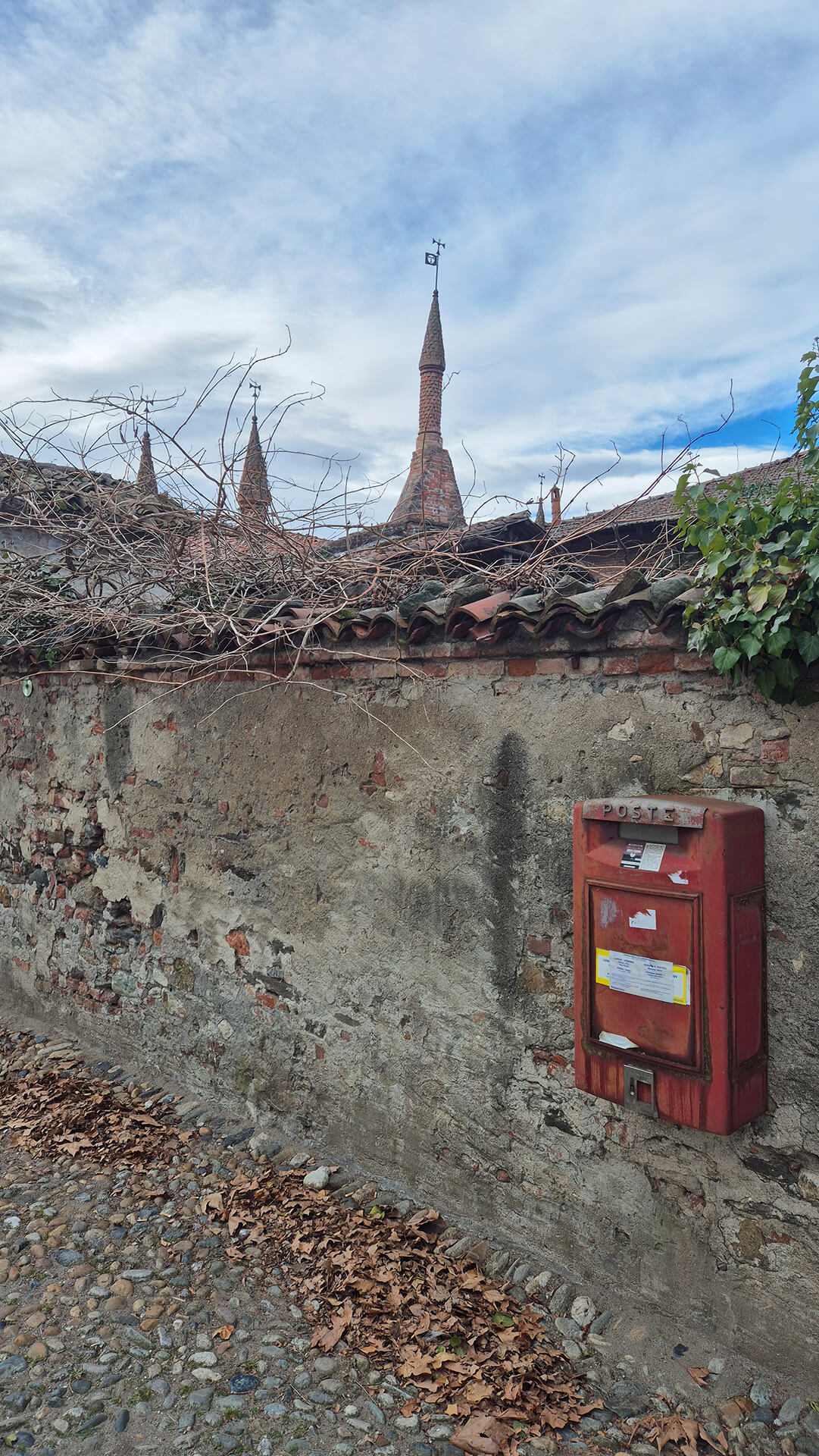 Fotografia di un muro perimetrale storico realizzato in muratura mista di mattoni e pietrame, caratterizzato da intonaco degradato e lacune che lasciano intravedere la tessitura originaria. La sommità del muro è protetta da una copertina in coppi di laterizio, sopra la quale si sviluppa vegetazione spontanea e rami secchi, segno di una manutenzione limitata e di un progressivo degrado superficiale. In primo piano è visibile una pavimentazione esterna in ciottoli con presenza di foglie secche, elemento che suggerisce un contesto di corte o di percorso storico pavimentato con materiali tradizionali. Sul lato destro del muro è fissata una cassetta postale metallica di colore rosso, con segni di usura e scolorimento, che testimonia l’adattamento contemporaneo di uno spazio antico a funzioni ancora in uso. Oltre il muro si intravedono coperture in coppi e alcune torrette in laterizio con forma conica e banderuola metallica, elementi architettonici che rimandano a un complesso storico di carattere religioso, rurale o fortificato. La presenza delle guglie e delle coperture tradizionali suggerisce un impianto edilizio di origine antica, probabilmente oggetto di trasformazioni nel tempo. L’immagine documenta lo stato materico e conservativo delle strutture murarie, mettendo in evidenza la stratificazione dei materiali, i segni del tempo e il rapporto tra elementi storici e inserimenti più recenti, utile per la lettura dello stato di fatto e per eventuali interventi di restauro o riqualificazione.