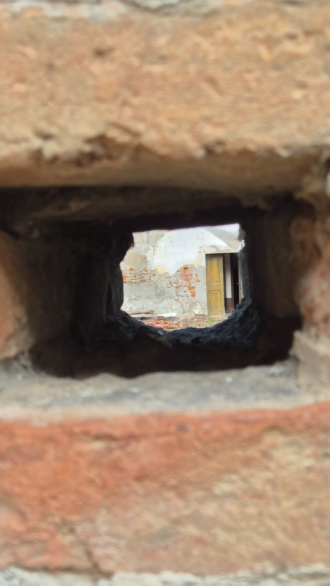 Dettaglio ravvicinato di un foro quadrangolare passante realizzato nella muratura in laterizio, probabilmente destinato in origine all’alloggiamento di travi di ponteggio o elementi strutturali temporanei utilizzati durante la costruzione dell’edificio. L’inquadratura è effettuata dall’esterno verso l’interno della cavità, mettendo in evidenza lo spessore della muratura e la finitura irregolare delle superfici interne, annerite e consumate, segno di lunga esposizione agli agenti atmosferici e di possibile utilizzo prolungato come sede di elementi lignei. Attraverso l’apertura è visibile uno scorcio della corte interna del complesso, con murature intonacate degradate, porzioni di laterizio a vista e una porta lignea, elementi che confermano la natura storica e stratificata dell’edificio. La presenza di lacune nell’intonaco e di parti murarie affioranti evidenzia fenomeni di distacco e usura, tipici di strutture non recentemente restaurate. I bordi del foro risultano irregolari e leggermente arrotondati, con fughe di malta erose e mattoni consumati, indicando un manufatto realizzato con tecnica tradizionale e non rifinito per essere a vista, ma destinato a una funzione costruttiva temporanea. Lo spessore consistente della parete suggerisce una muratura portante di notevole massa, coerente con edifici di carattere rurale, conventuale o di servizio. L’immagine è utile per la lettura del dettaglio costruttivo della muratura, delle tecniche di cantiere storiche e dello stato di conservazione dei materiali, fornendo indicazioni importanti per analisi stratigrafiche, rilievi architettonici e valutazioni di intervento conservativo.