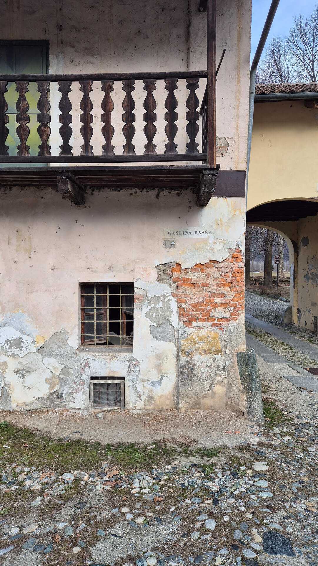 Vista frontale di una porzione di edificio rurale storico con ballatoio ligneo, appartenente al complesso edilizio già documentato, caratterizzato da muratura portante intonacata con ampie lacune che lasciano a vista il laterizio sottostante. Al piano superiore è presente un balcone in legno con parapetto sagomato, sostenuto da mensole lignee sporgenti, tipico delle architetture agricole tradizionali dell’area piemontese, probabilmente utilizzato in origine come spazio di servizio o di collegamento tra ambienti del piano primo. La superficie muraria mostra evidenti fenomeni di degrado, con distacchi diffusi dell’intonaco, efflorescenze, macchie di umidità di risalita e porzioni di muratura in laterizio irregolare affiorante, che consentono di leggere la tecnica costruttiva originaria. Al piano terra si apre una finestra di piccole dimensioni con grata metallica, affiancata da una bocca di aerazione o apertura di ventilazione posta in basso, elementi che suggeriscono la presenza di locali di servizio o seminterrati. Sulla parete è visibile la scritta “CASCINA BASSA”, indicazione toponomastica che conferma la natura rurale del complesso e la sua articolazione in più corpi di fabbrica con denominazioni specifiche. Sul lato destro si apre un passaggio voltato ad arco ribassato, che mette in comunicazione la corte interna con lo spazio esterno, definendo un sistema distributivo tipico delle cascine storiche, organizzate attorno a cortili chiusi. La pavimentazione antistante è costituita da terreno battuto e ciottoli, in continuità con le sistemazioni tradizionali della corte, mentre lo stato di conservazione generale evidenzia un edificio non recentemente restaurato, ma ancora leggibile nelle sue caratteristiche originarie. L’immagine è significativa per l’analisi del corpo rurale secondario, delle tecniche costruttive tradizionali, dello stato di degrado dell’intonaco e dell’organizzazione della cascina attorno a spazi di corte e passaggi voltati, elementi utili per rilievo architettonico, studio storico-tipologico e valutazioni di recupero conservativo.