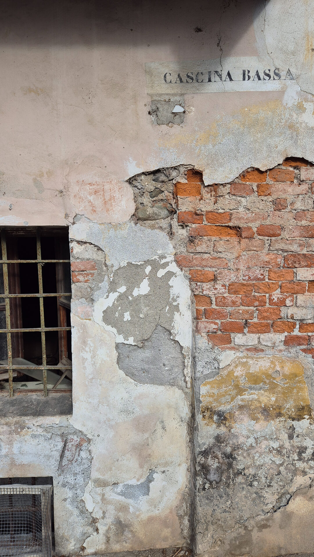 Dettaglio ravvicinato della muratura intonacata appartenente al corpo edilizio denominato “Cascina Bassa”, con evidenza della scritta toponomastica realizzata a stencil direttamente sull’intonaco, parzialmente deteriorato. La superficie muraria mostra un avanzato stato di degrado, con ampie lacune dell’intonaco che lasciano a vista la struttura sottostante in laterizio pieno disposto a corsi irregolari, legato con malta di allettamento ormai erosa in più punti. Nella parte sinistra è presente una finestra di piccole dimensioni con inferriata metallica, inserita in una spalletta intonacata fortemente deteriorata, con distacchi, cavillature e zone di rappezzo realizzate con malte di diversa composizione e granulometria. Al di sotto della finestra si riconosce una piccola apertura di aerazione protetta da rete metallica, indicativa della presenza di locali seminterrati o di servizio, coerente con la tipologia costruttiva delle cascine tradizionali. La muratura presenta evidenti fenomeni di umidità di risalita capillare, riconoscibili dalle macchie scure, dalle efflorescenze e dal disgregamento dell’intonaco nella fascia inferiore, oltre a depositi superficiali e alterazioni cromatiche che indicano esposizione prolungata agli agenti atmosferici. La discontinuità tra le parti intonacate e quelle in laterizio a vista consente di leggere chiaramente la stratificazione degli interventi manutentivi succedutisi nel tempo. La presenza della scritta identificativa “CASCINA BASSA” conferma l’appartenenza dell’edificio al sistema della cascina storica già documentata nelle immagini precedenti, evidenziando il carattere rurale del complesso e la sua articolazione in più corpi di fabbrica. L’immagine risulta utile per l’analisi dello stato di conservazione delle superfici murarie, dei fenomeni di degrado dell’intonaco e delle tecniche costruttive tradizionali in laterizio, fornendo indicazioni rilevanti per rilievo materico, diagnosi del degrado e valutazioni di intervento conservativo.
