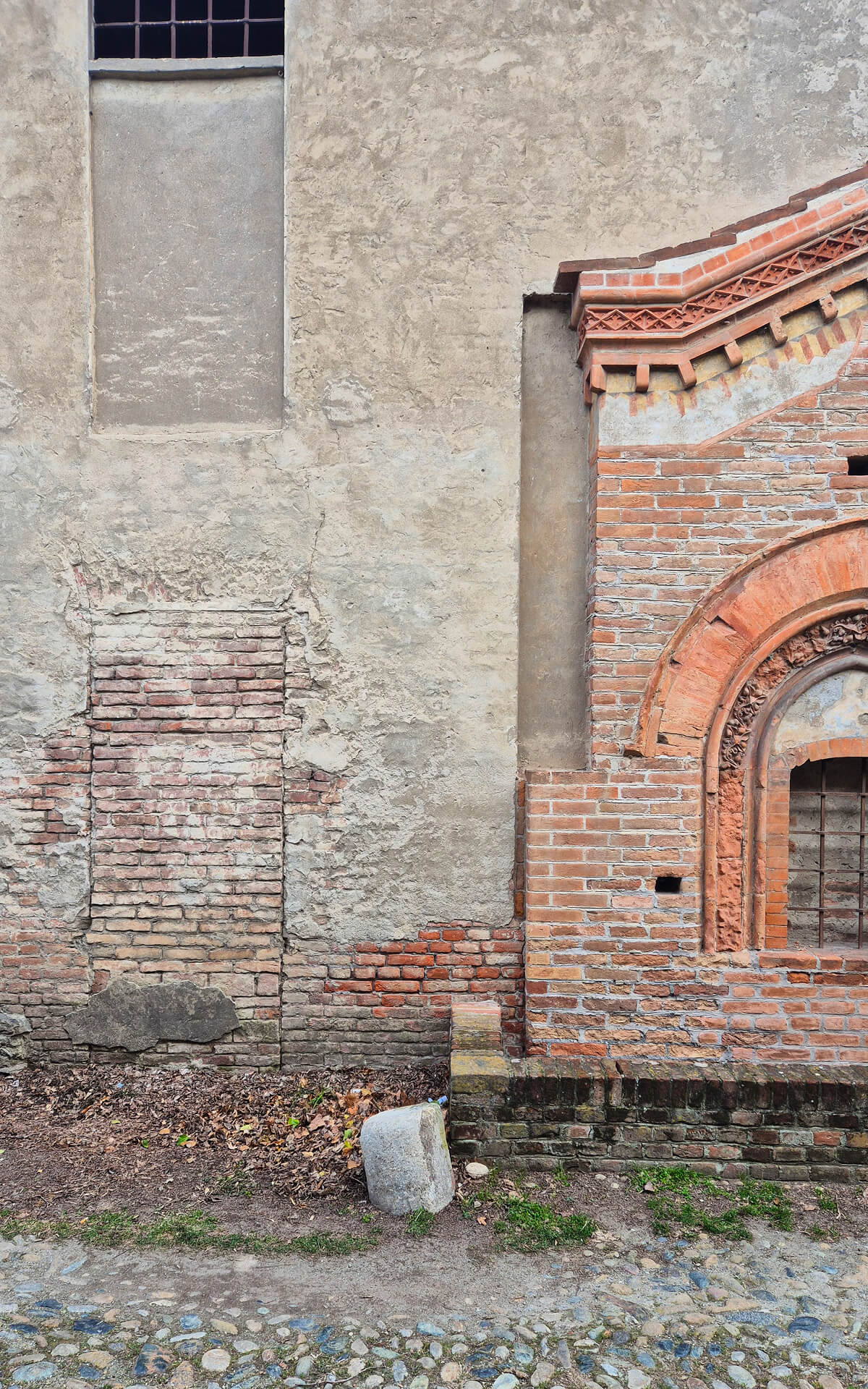 ettaglio ravvicinato di una muratura storica che evidenzia la stratificazione costruttiva e lo stato materico di una facciata in laterizio e intonaco. L’immagine mostra l’incontro tra due corpi edilizi differenti: sulla sinistra una parete intonacata fortemente degradata, con ampie porzioni di rivestimento cadute che lasciano scoperta la tessitura in mattoni, e sulla destra un elemento architettonico in laterizio faccia a vista con decorazioni di gusto gotico. La parte in mattoni presenta una piccola apertura ad arco acuto incorniciata da elementi sagomati in cotto, con motivo ornamentale nella ghiera e cornice superiore decorata con fasce dentellate e modanature. Questo apparato decorativo, realizzato con grande cura artigianale, contrasta con la superficie intonacata adiacente, più povera e segnata da interventi successivi, tamponamenti e riparazioni che testimoniano modifiche nel tempo. Nella porzione sinistra si osserva chiaramente una tamponatura in laterizio di una precedente apertura, riconoscibile dalla differenza di tessitura e dalla forma rettangolare inserita nella muratura. La presenza di crepe, distacchi di intonaco, efflorescenze e discontinuità cromatiche indica un avanzato stato di degrado superficiale, utile per la lettura delle fasi costruttive e degli interventi successivi. Alla base della parete è visibile il contatto con il terreno e con una pavimentazione esterna in ciottoli, mentre piccoli elementi lapidei e accumuli di foglie segnalano uno spazio esterno poco manutenuto. L’immagine ha valore documentario per l’analisi dello stato di fatto, mettendo in evidenza materiali, tecniche costruttive, stratificazioni e condizioni conservative utili in vista di un eventuale intervento di restauro.