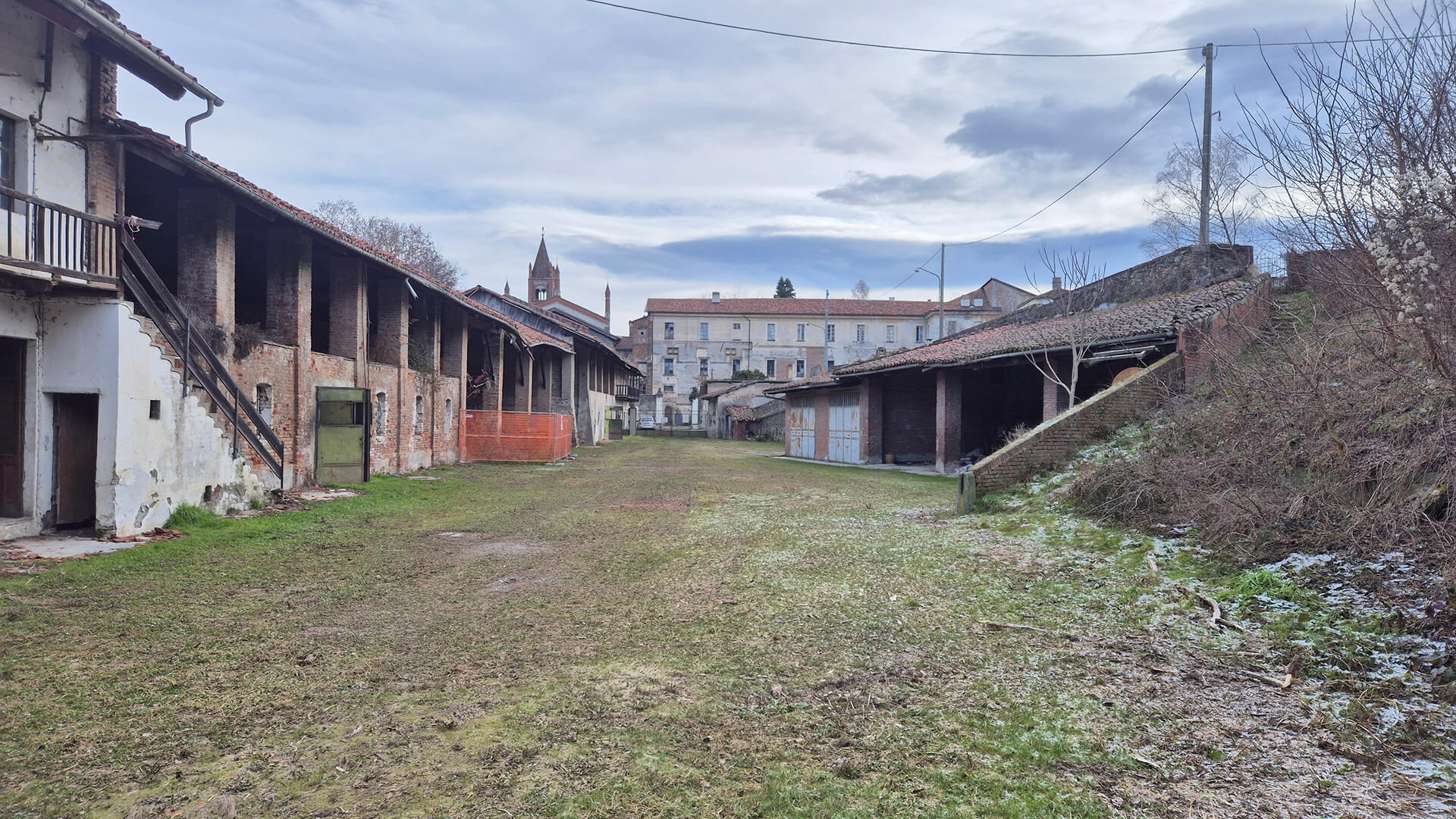 Vista d’insieme di uno spazio aperto interno a un complesso rurale storico, configurato come corte rettangolare delimitata da edifici porticati e fabbricati di servizio, realizzati prevalentemente in muratura mista di laterizio e pietrame con coperture a falde in coppi. L’immagine mostra chiaramente l’impianto tipico di cascina o complesso agricolo, con corpi di fabbrica disposti sui lati lunghi della corte e spazi aperti destinati originariamente alle attività produttive e di servizio. Sul lato sinistro si sviluppa un edificio a due livelli con loggiato continuo al piano superiore, sostenuto da pilastri in muratura, e scala esterna di accesso in ferro e legno. Le murature presentano ampie porzioni di intonaco degradato che lasciano a vista il laterizio, evidenziando lo stato di conservazione non omogeneo e la presenza di interventi di manutenzione parziale. Al piano terra si aprono vani di servizio e accessi carrabili, alcuni chiusi con serramenti metallici. Sul lato destro è presente un corpo più basso, caratterizzato da porticati aperti e autorimesse o depositi, con copertura inclinata e struttura semplice, probabilmente destinato a ricovero attrezzi o stalle. La continuità dei fronti e la disposizione parallela degli edifici definiscono uno spazio centrale ampio, oggi a prato, che conserva la funzione di area di manovra e distribuzione. Il terreno della corte è in parte erboso e in parte sterrato, con segni di umidità e scarsa manutenzione, mentre sul fondo si intravede un edificio di maggior pregio architettonico, probabilmente residenziale o istituzionale, che chiude prospetticamente lo spazio. Sullo sfondo emerge inoltre un campanile in laterizio, elemento che colloca il complesso in un contesto storico stratificato di ambito rurale o periurbano. L’immagine è significativa per la lettura dell’impianto tipologico originario, dello stato di conservazione e delle trasformazioni funzionali del complesso, utile per analisi preliminari di rilievo, restauro o progetto di rifunzionalizzazione.