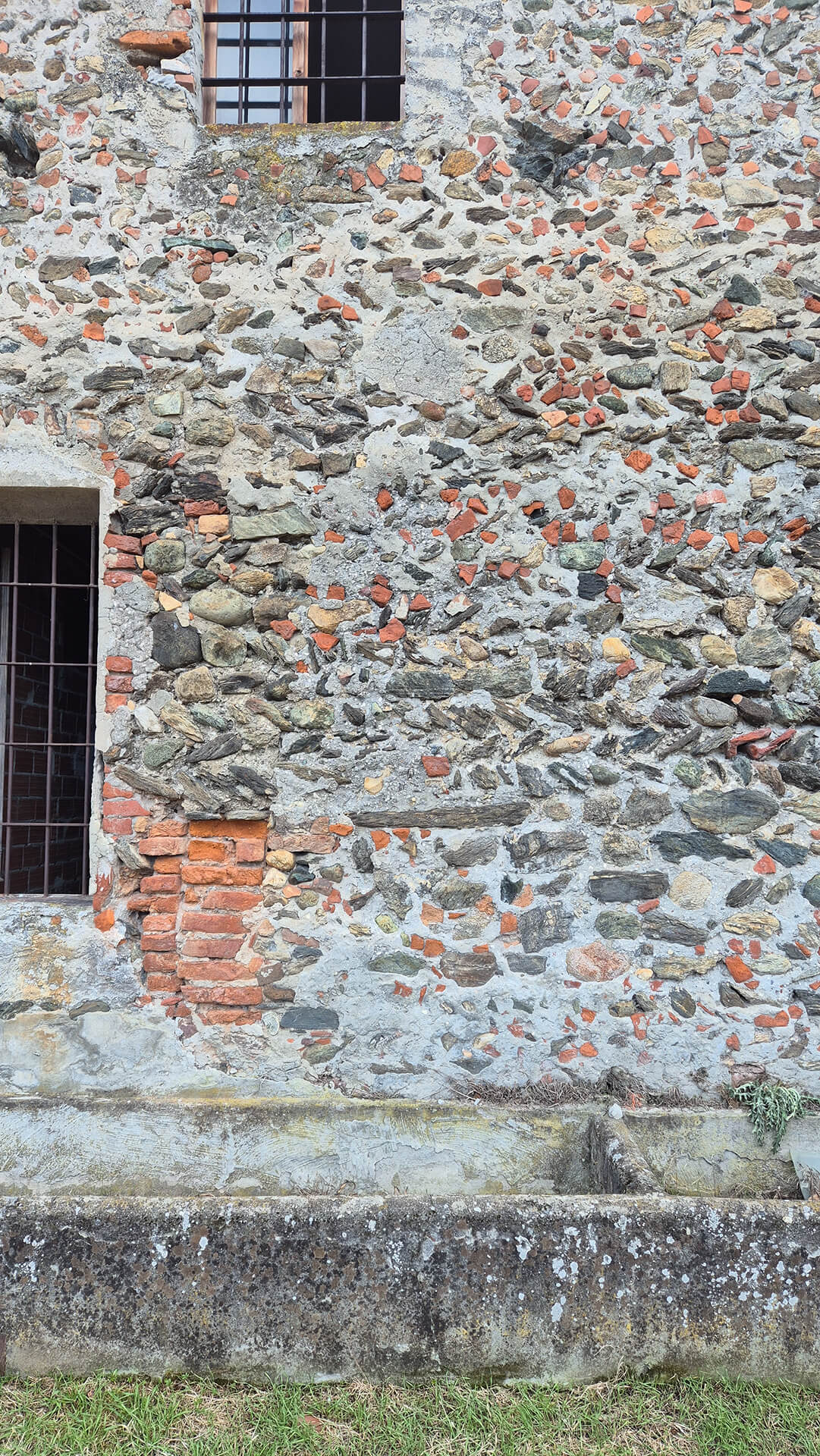 Dettaglio di facciata in muratura mista storica composta da pietrame, ciottoli fluviali e frammenti di laterizio, legati con malta di calce, caratterizzata da tessitura irregolare e forte eterogeneità materica. Il paramento mostra chiaramente una tecnica costruttiva povera, realizzata con materiali di recupero disposti senza ordine regolare, con giunti ampi e non uniformi, tipica degli edifici rurali e di servizio. Sono visibili due aperture sovrapposte con inferriate metalliche, inserite in vani con contorni in laterizio e pietra, che evidenziano adattamenti successivi rispetto alla muratura originaria. Attorno alla finestra inferiore si notano integrazioni in mattoni pieni disposti con maggiore regolarità, probabilmente dovute a interventi di consolidamento o alla modifica della dimensione del vano. La differenza di colore e di tessitura tra i laterizi e il resto della parete permette di leggere chiaramente le diverse fasi costruttive. La superficie muraria presenta diffusi fenomeni di degrado, tra cui erosione dei giunti, distacchi di malta, discontinuità cromatiche e piccole fessurazioni, dovuti all’esposizione agli agenti atmosferici e alla presenza di umidità. Alla base della parete è presente una canaletta di drenaggio in calcestruzzo, addossata al piede della muratura, con funzione di raccolta delle acque meteoriche e di protezione del basamento dall’umidità di ristagno. Anche in questa zona si osservano depositi, colonizzazione biologica e alterazioni superficiali. L’immagine è significativa per l’analisi dello stato di conservazione, della tecnica muraria tradizionale e delle trasformazioni subite nel tempo, mettendo in evidenza stratificazioni, tamponamenti e interventi successivi, elementi fondamentali per la lettura storico-costruttiva e per eventuali interventi di restauro o consolidamento.