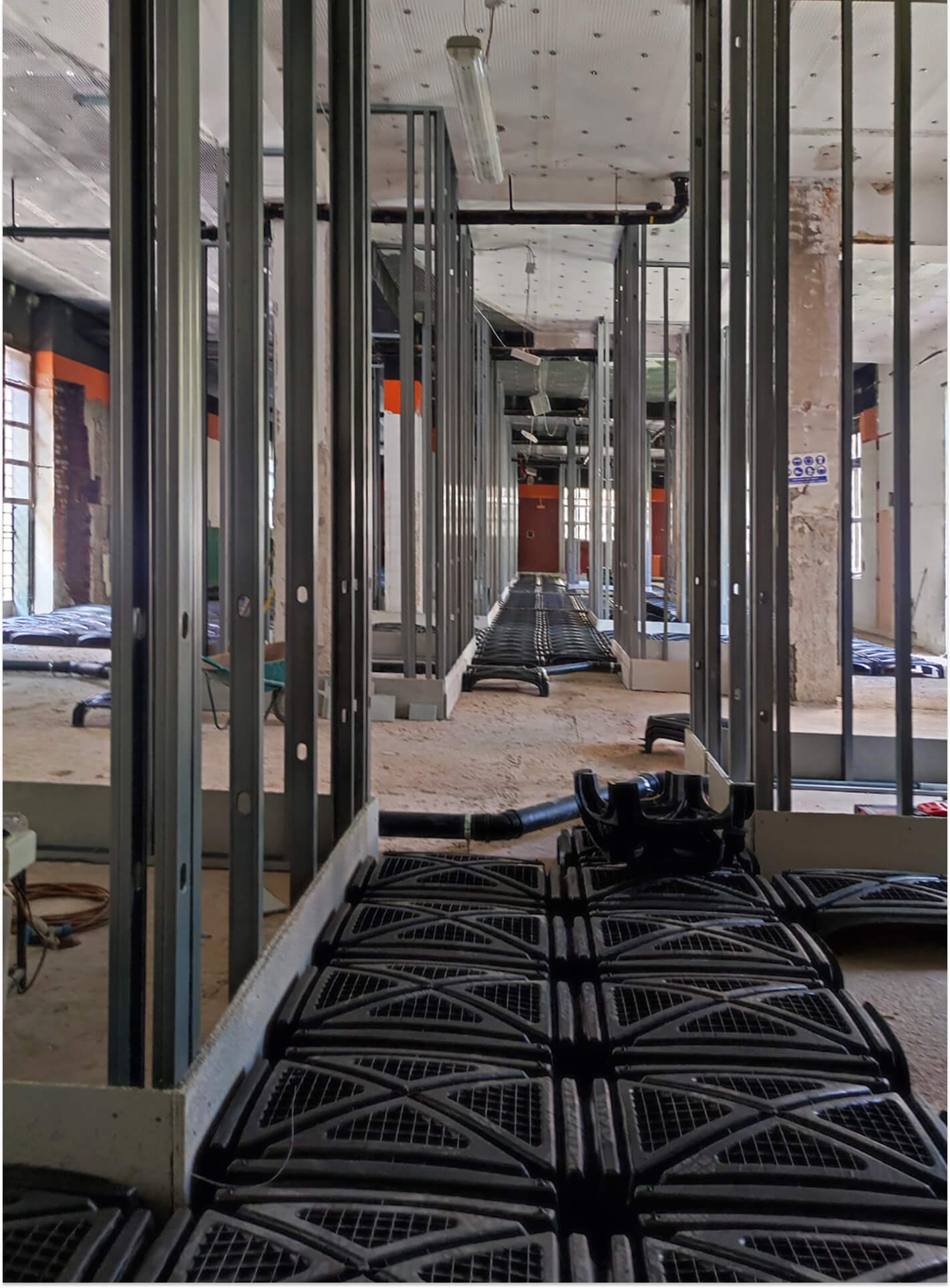 Vista interna di ambiente in fase di riqualificazione, con realizzazione di nuove partizioni distributive mediante struttura metallica leggera per sistemi a secco. L’immagine mostra l’orditura in profili zincati fissati a pavimento e a soffitto, predisposta per la successiva chiusura con pannelli in cartongesso, mentre al suolo sono visibili moduli plastici per la formazione di vespaio aerato e canalizzazioni impiantistiche integrate nel massetto. Lungo l’asse centrale si sviluppa il tracciato degli impianti, con tubazioni e passaggi tecnici posati prima della realizzazione del sottofondo, evidenziando la fase di coordinamento tra opere edili e impiantistiche. I pilastri esistenti in calcestruzzo armato rimangono a vista e vengono inglobati nel nuovo assetto distributivo, mentre il controsoffitto tecnico consente il passaggio delle reti impiantistiche orizzontali. La fotografia documenta una fase intermedia di cantiere, in cui sono leggibili le stratigrafie costruttive e le soluzioni adottate per l’adeguamento funzionale e tecnologico dell’edificio esistente.