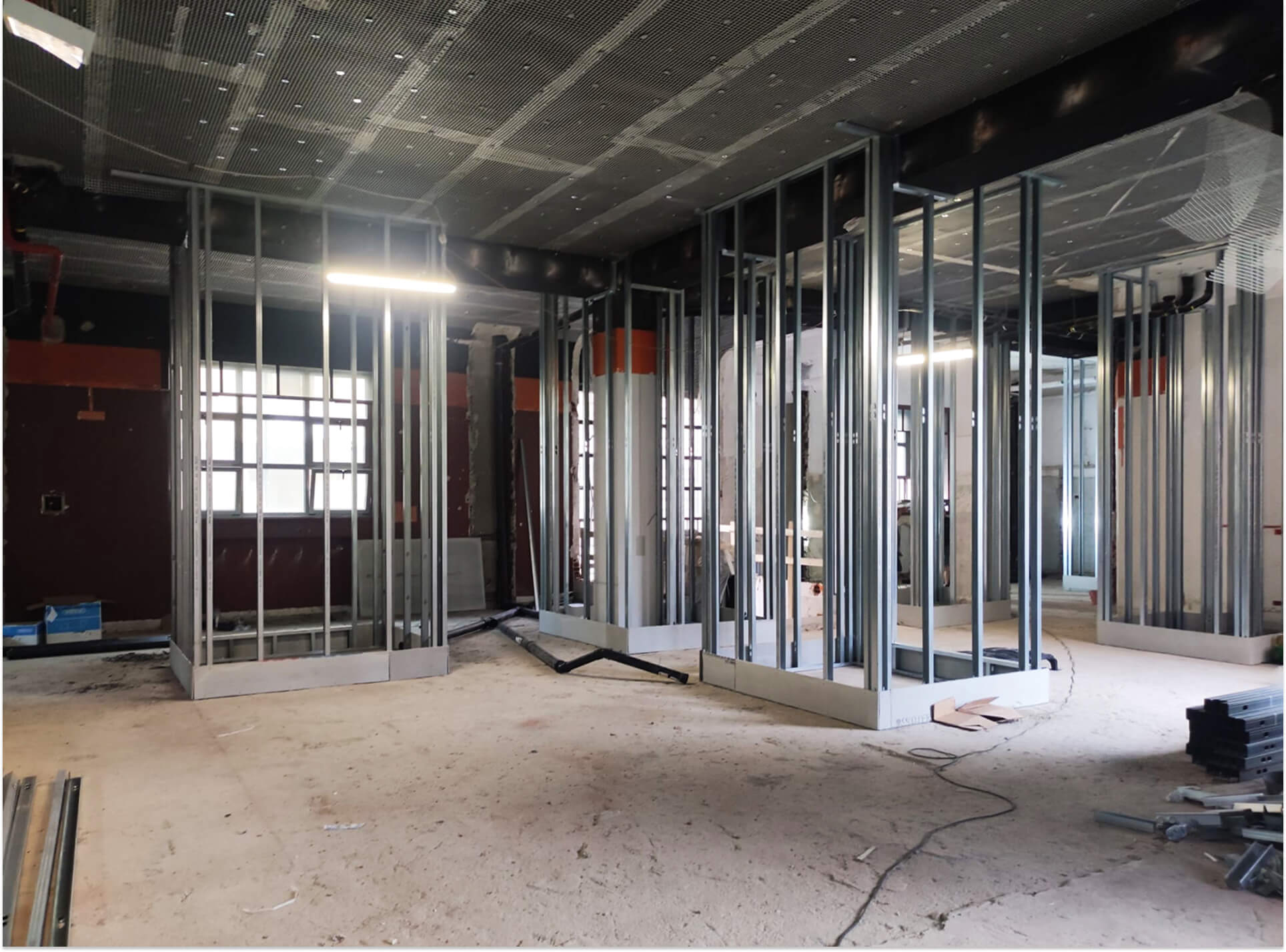 Vista interna di ambiente in fase di ristrutturazione, con configurazione spaziale in corso di definizione mediante realizzazione di nuove partizioni con sistema a secco su struttura metallica leggera. Sono visibili i telai in profili di acciaio zincato ancorati al solaio e al pavimento, predisposti per la successiva chiusura con pannelli in cartongesso, che delineano i nuovi locali secondo il progetto distributivo. Il soffitto è configurato come controsoffitto tecnico continuo, con pannellatura microforata e struttura sospesa che consente l’alloggiamento degli impianti, tra cui canalizzazioni, tubazioni e corpi illuminanti temporanei di cantiere. I pilastri strutturali esistenti in calcestruzzo armato rimangono a vista e vengono integrati nel nuovo assetto interno, mentre il pavimento è ancora allo stato grezzo, in attesa della realizzazione dei sottofondi e delle finiture. L’immagine documenta una fase intermedia dei lavori, evidenziando il coordinamento tra opere edilizie, sistemi a secco e adeguamento impiantistico nell’ambito della rifunzionalizzazione dell’edificio.