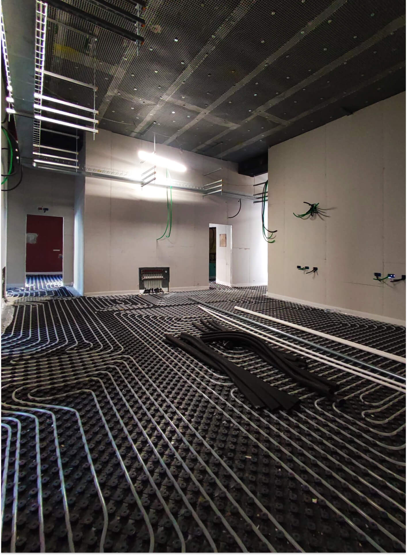 Ambiente interno in fase avanzata di riqualificazione impiantistica, con predisposizione del sistema di riscaldamento a pavimento mediante posa di pannelli modulari bugnati e serpentine in tubazione plastica per impianto radiante annegato nel massetto. La distribuzione delle linee segue un tracciato regolare su tutta la superficie del locale, con collettore di distribuzione installato a parete e tubazioni di adduzione ancora in fase di collegamento. Le pareti perimetrali e divisorie risultano già chiuse con pannelli in cartongesso su struttura metallica, mentre il controsoffitto tecnico sospeso con pannellatura microforata consente l’alloggiamento delle canalizzazioni impiantistiche, delle passerelle portacavi e dei corpi illuminanti temporanei di cantiere. Sono visibili le predisposizioni per impianti elettrici e speciali con cavidotti emergenti dalle pareti e dal soffitto. Il pavimento si trova nella fase precedente alla realizzazione del massetto di finitura che ingloberà l’impianto radiante, documentando una fase intermedia di cantiere in cui risultano coordinate le lavorazioni edilizie e impiantistiche finalizzate all’adeguamento tecnologico e funzionale dell’edificio.