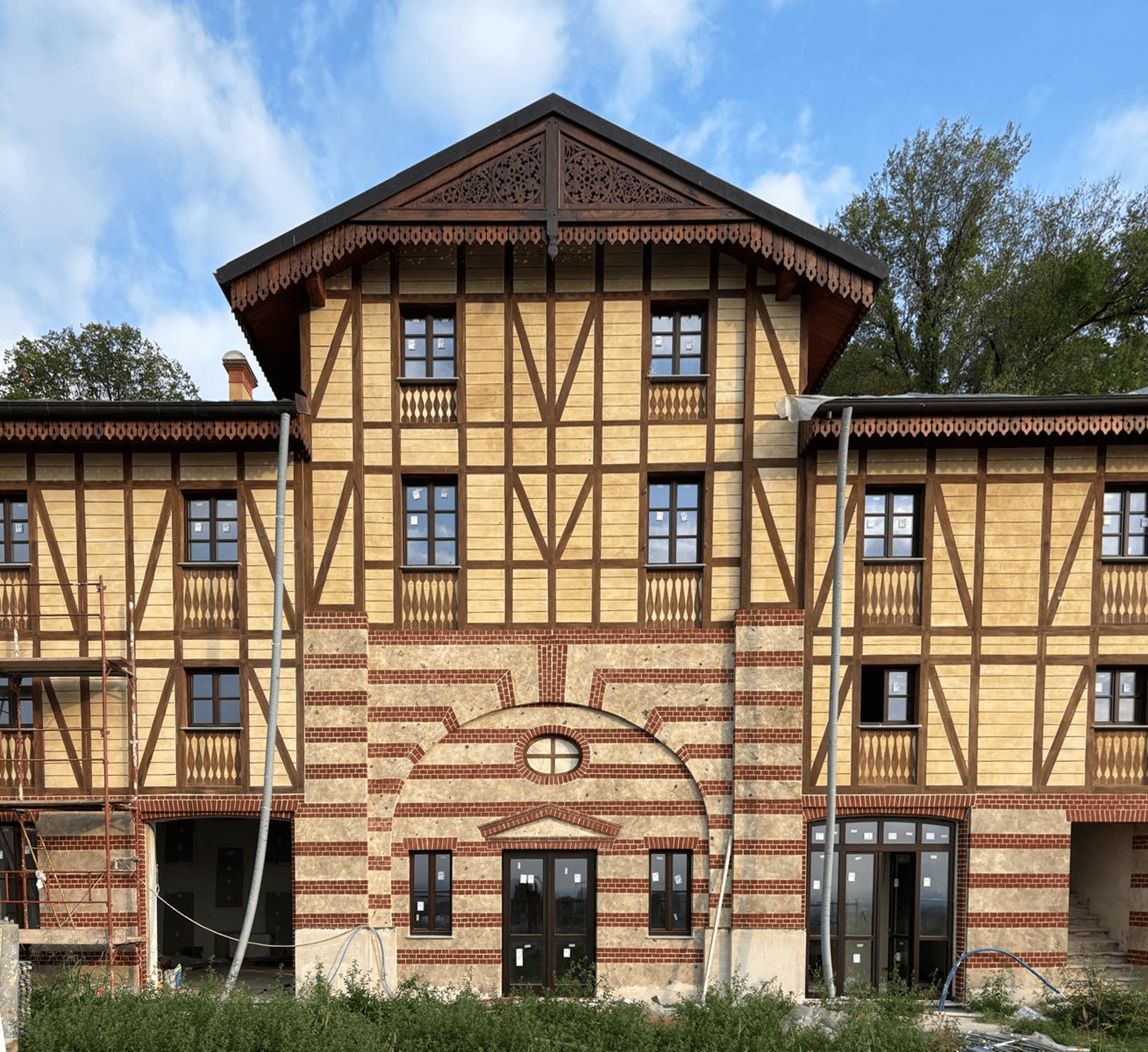 L'immagine mostra una facciata di un edificio in fase di ristrutturazione, probabilmente un esempio di restauro di una struttura storica. Ecco alcuni dettagli evidenti: Stile architettonico: L'edificio presenta uno stile tradizionale che richiama l'architettura half-timbered (a traliccio di legno), con un mix di legno e mattoni, tipico di alcune zone dell'Europa, come la Francia e la Germania. Le travi di legno in rilievo contrastano con le pareti in muratura, creando un effetto visivo distintivo. Materiali e finiture: La struttura combina il legno chiaro con mattoni rossi, che decorano la base dell'edificio, evidenziando le finestre e le porte. La parte superiore della facciata è decorata con un bordo intagliato di legno, che aggiunge un tocco di eleganza e dettaglio artigianale. La presenza di una scalinata di impalcature suggerisce che il lavoro di ristrutturazione è ancora in corso. Finestrature: Le finestre sono incorniciate da travi di legno scuro, con un design semplice ma elegante, e sono ancora protette con pellicole di plastica che indicano che non sono ancora pronte per l'uso. Spazio verde: Davanti all'edificio, c'è una piccola area di terreno verde che appare incolta, ma probabilmente sarà riorganizzata o rifinita una volta completata la ristrutturazione. Questo edificio potrebbe essere parte di un progetto di recupero di un'area storica, volto a preservare il fascino tradizionale mentre si integrano soluzioni moderne per la funzionalità e la vivibilità.